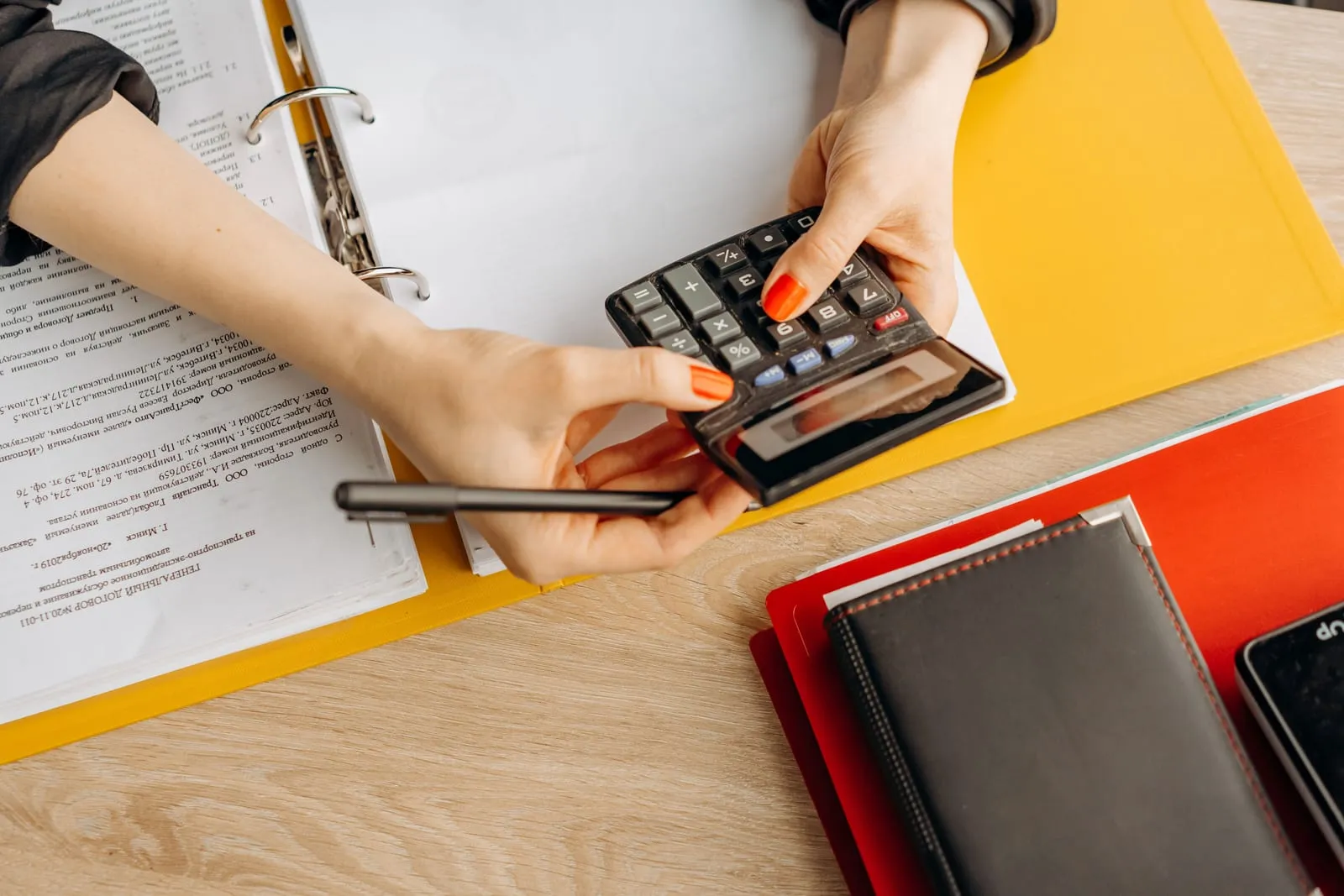 Hands with orange nails hold a desk calculator and a pen above an open yellow ring binder of printed documents, with a red folder and notebook nearby.