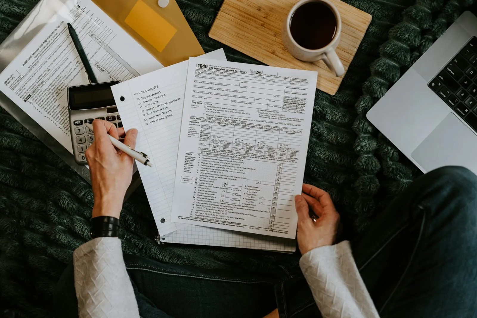 Overhead view of a person filling out a Form 1040 tax return with a pen, a handwritten tax documents checklist, a calculator, a laptop and a cup of coffee.