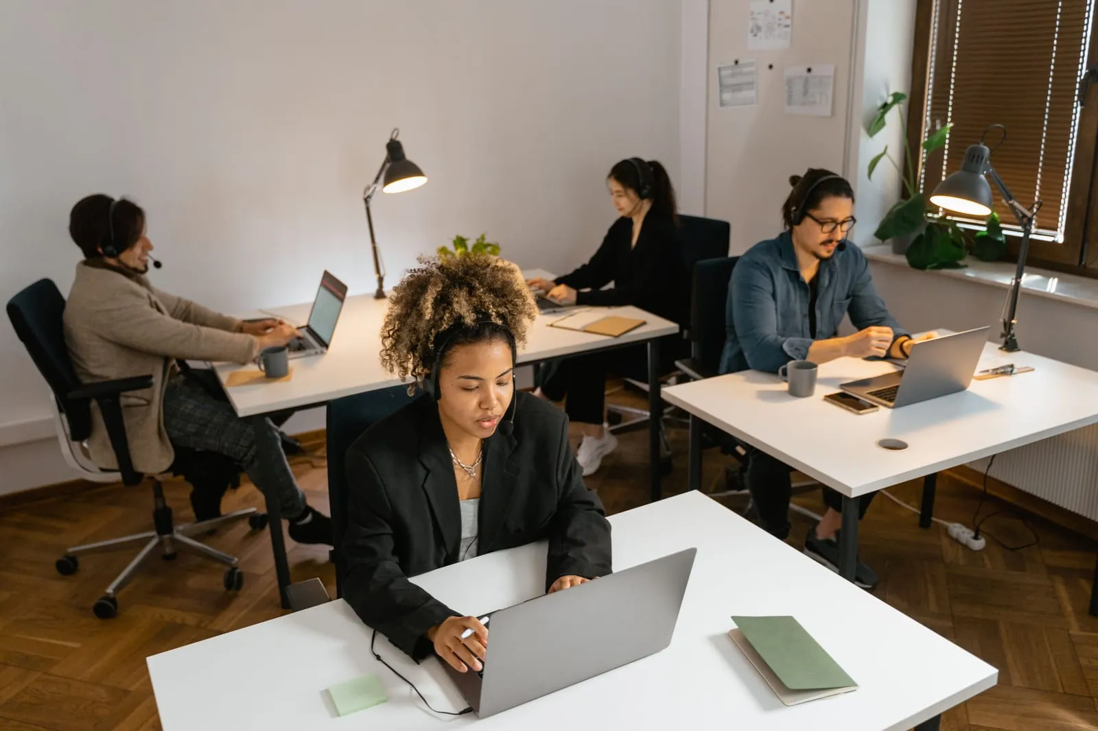 Four remote-style workers in headsets type on laptops at separate white desks in a bright office with desk lamps and parquet flooring.