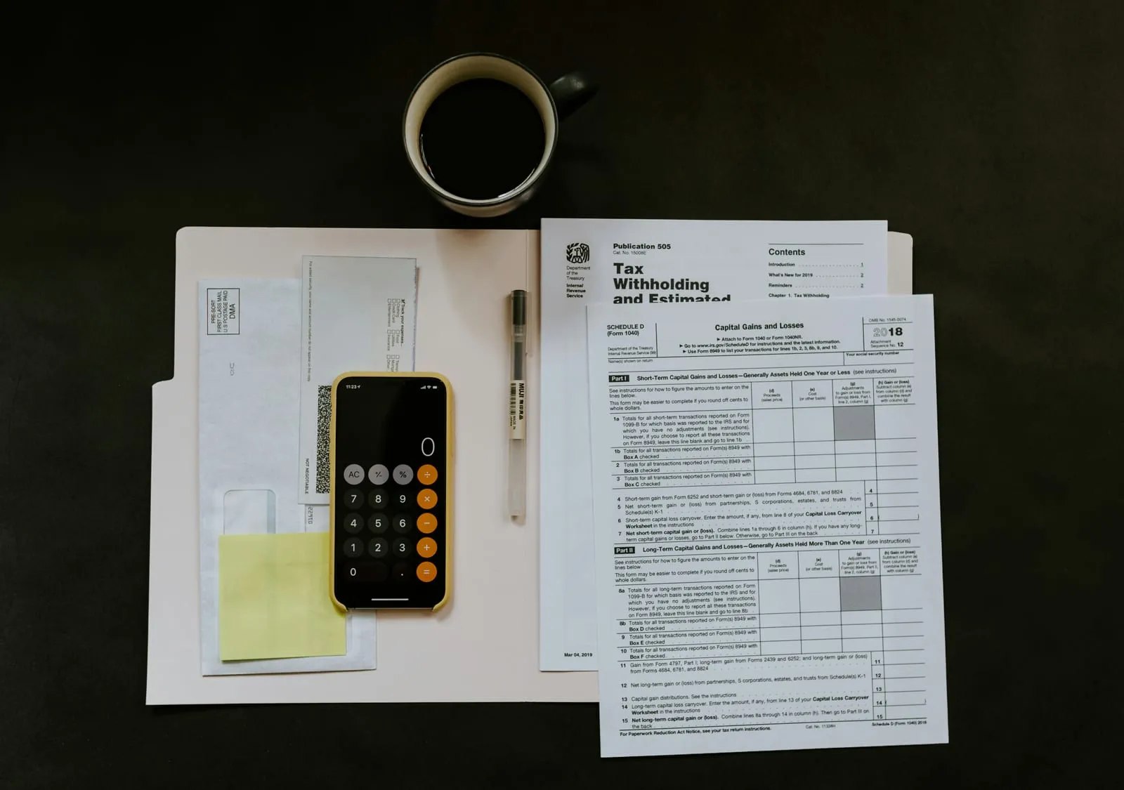Flat lay on a dark desk of IRS tax forms, a pale folder with envelopes, a phone showing a calculator, a pen and a black coffee mug.