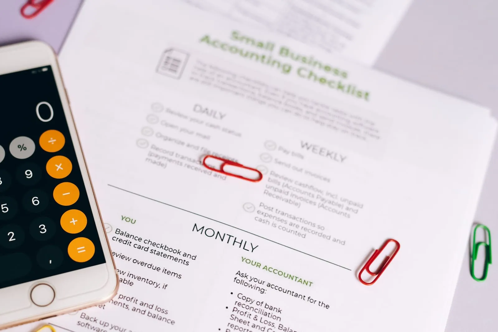 A printed small business accounting checklist with daily, weekly and monthly tasks, held down by red paper clips beside a smartphone calculator.