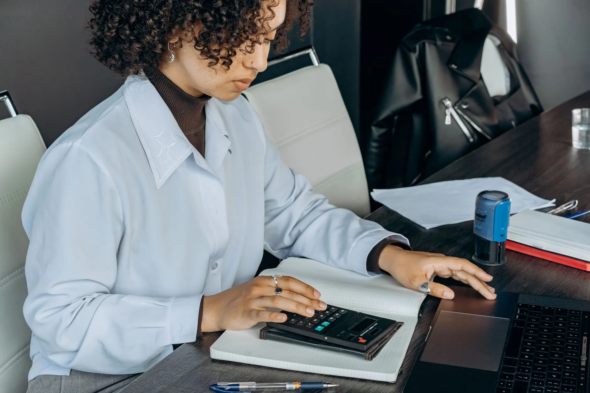 A woman in a pale blue shirt works a desktop calculator over a squared notebook at a dark wood desk, with a laptop, loose papers and a blue self-inking stamp beside her.