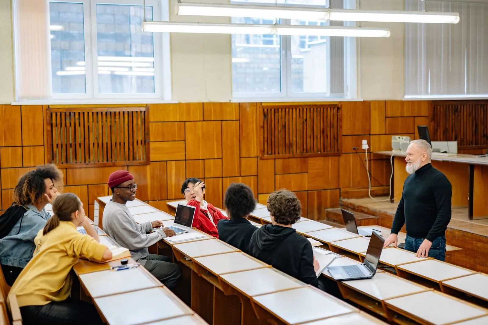 Gray-bearded instructor stands at the side of a tiered lecture hall chatting with students who have laptops and notebooks at wooden desks