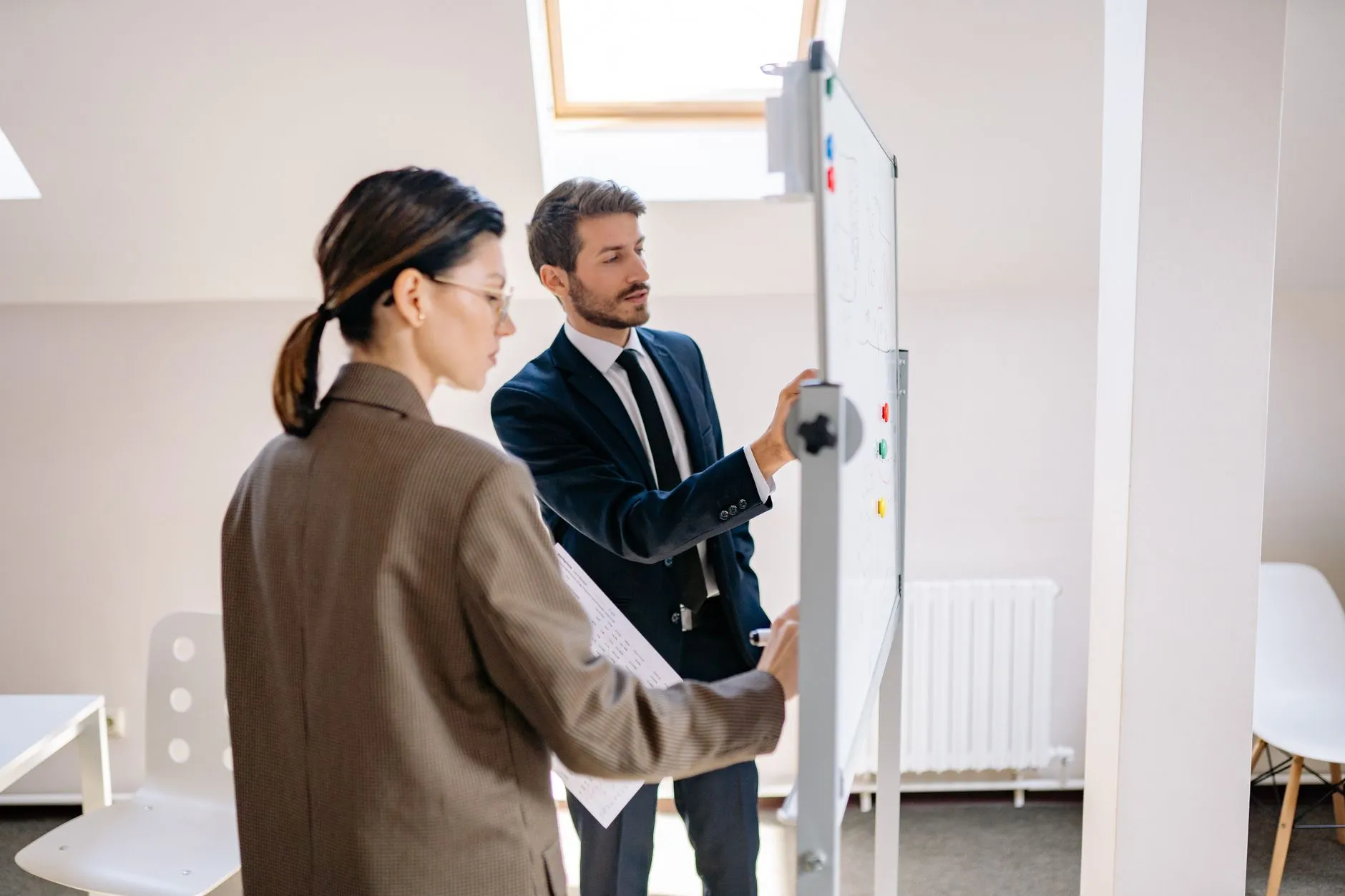 Two colleagues in business suits work through a plan on a rolling whiteboard in a bright office, one pointing at the board while the other holds printed notes.