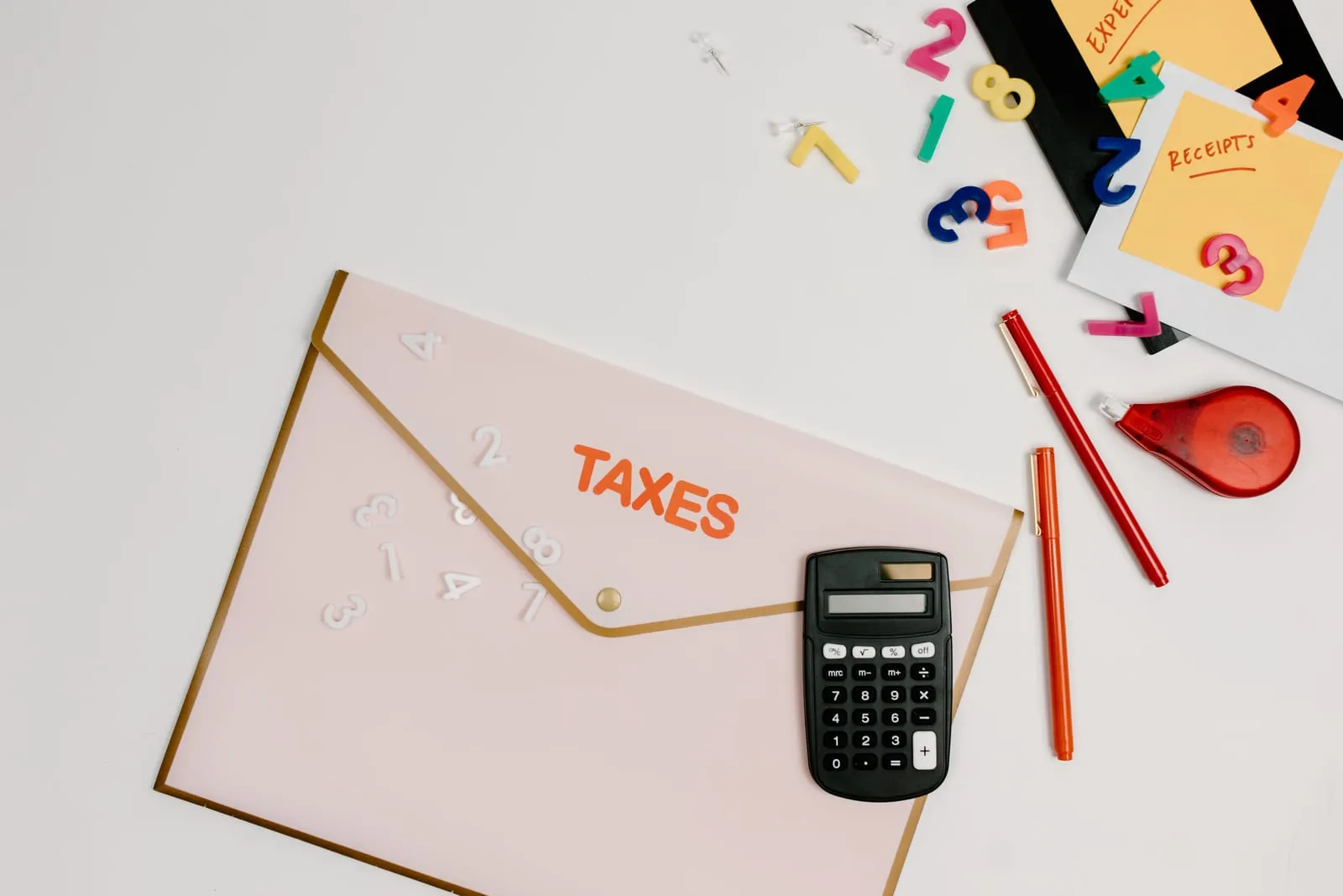 Flat lay of a pink tax folder, a calculator, red pens and colorful number magnets on a white surface
