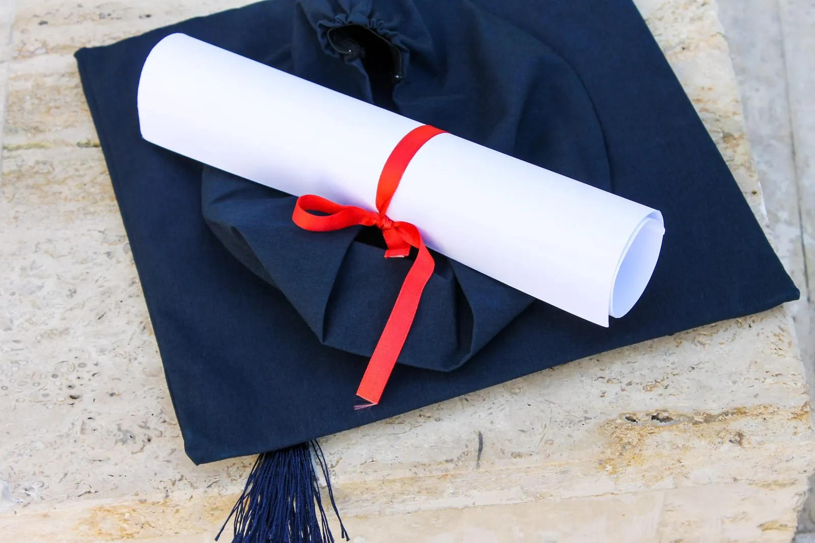 Rolled diploma tied with a red ribbon lying on a navy graduation cap on a stone ledge