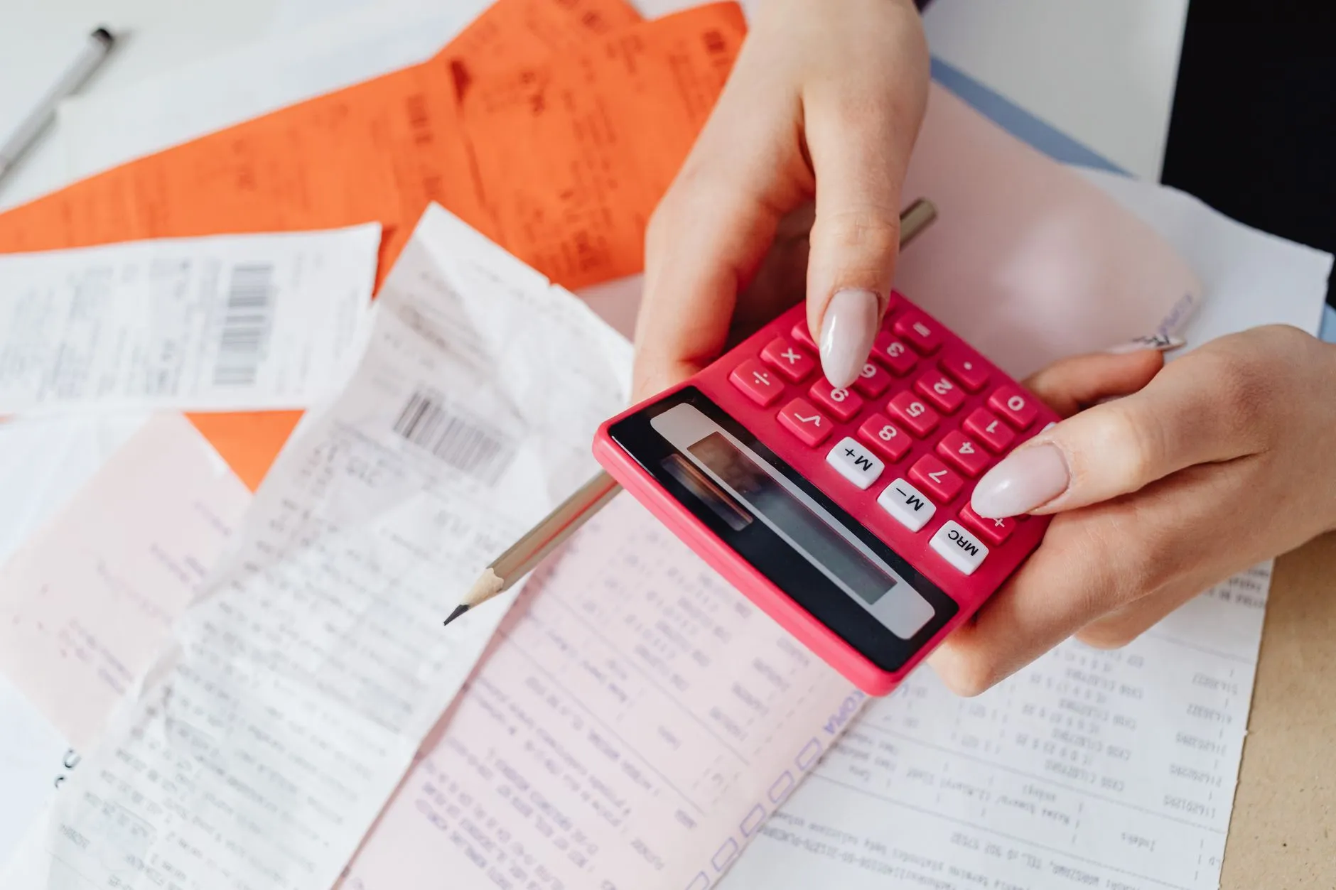 Hands holding a bright pink calculator over a scatter of printed receipts, with a pencil resting across the paper