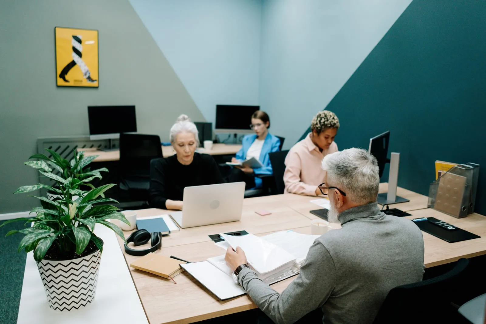 Four coworkers at shared desks in a teal and gray office, one older man in the foreground paging through a stack of documents