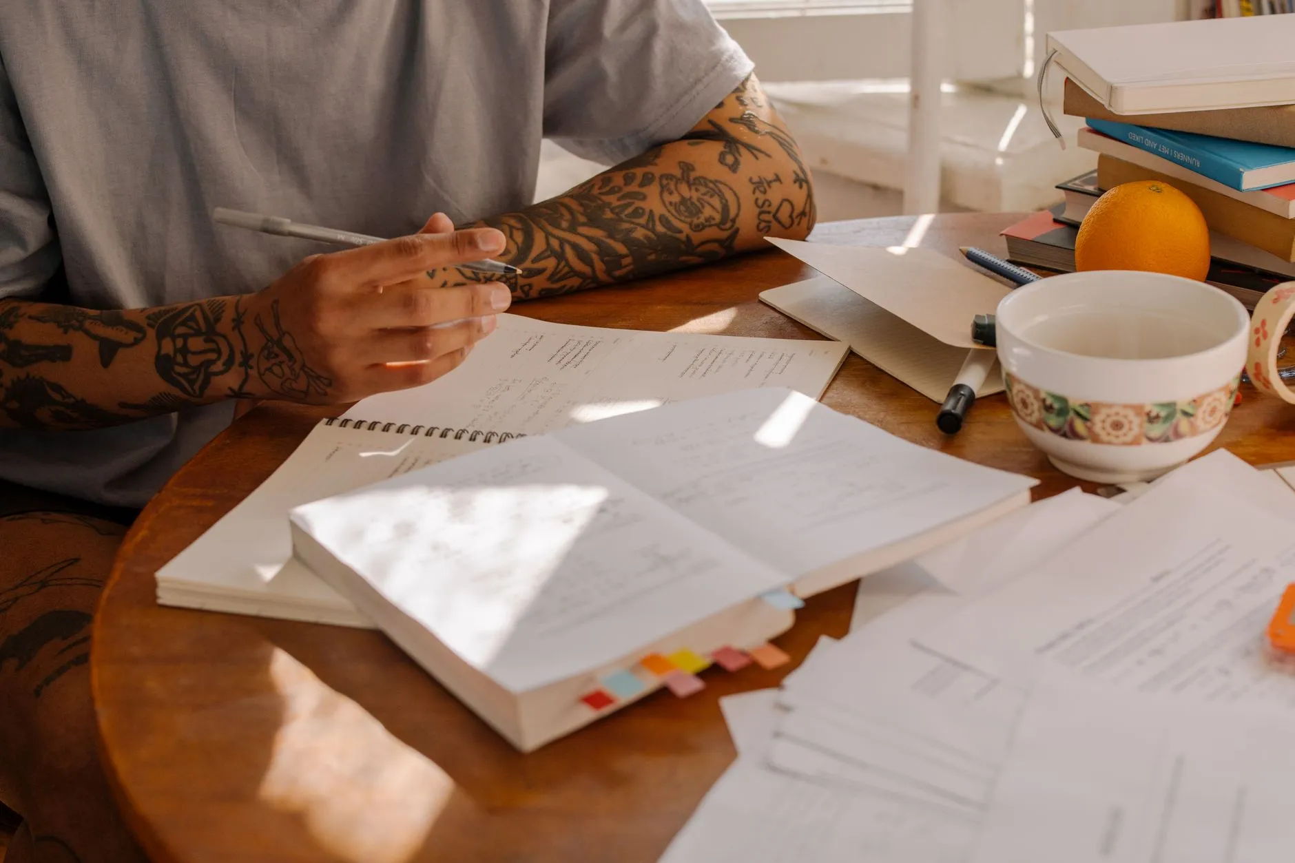 A person working through a tabbed study guide at a sunlit wooden table, with handwritten notes, loose practice sheets, a stack of books and a coffee bowl.