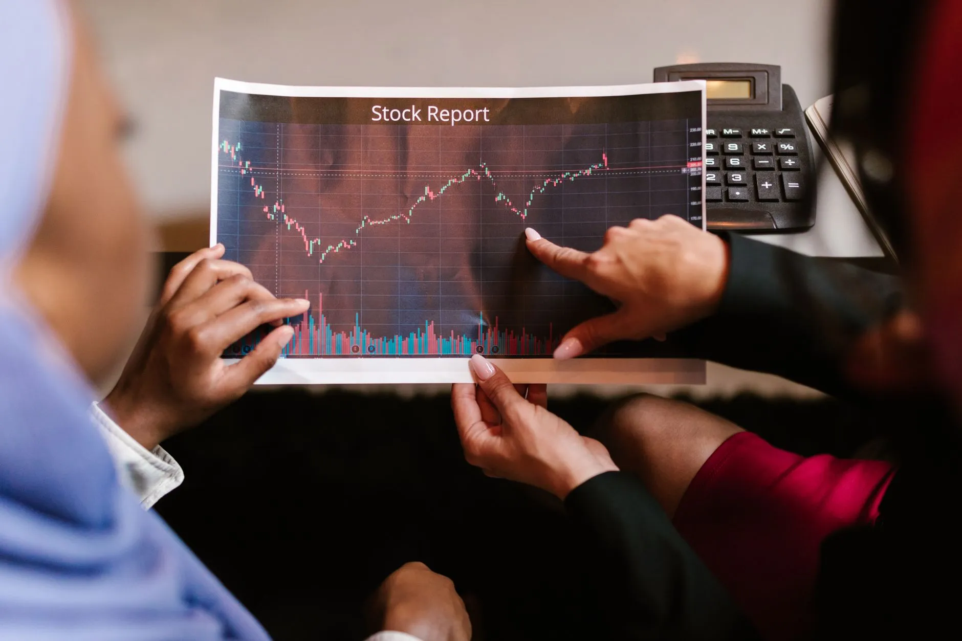 Two colleagues hold a printed stock report showing a candlestick price chart and trading volume bars; one points at a dip in the line while a desk calculator sits behind the page.