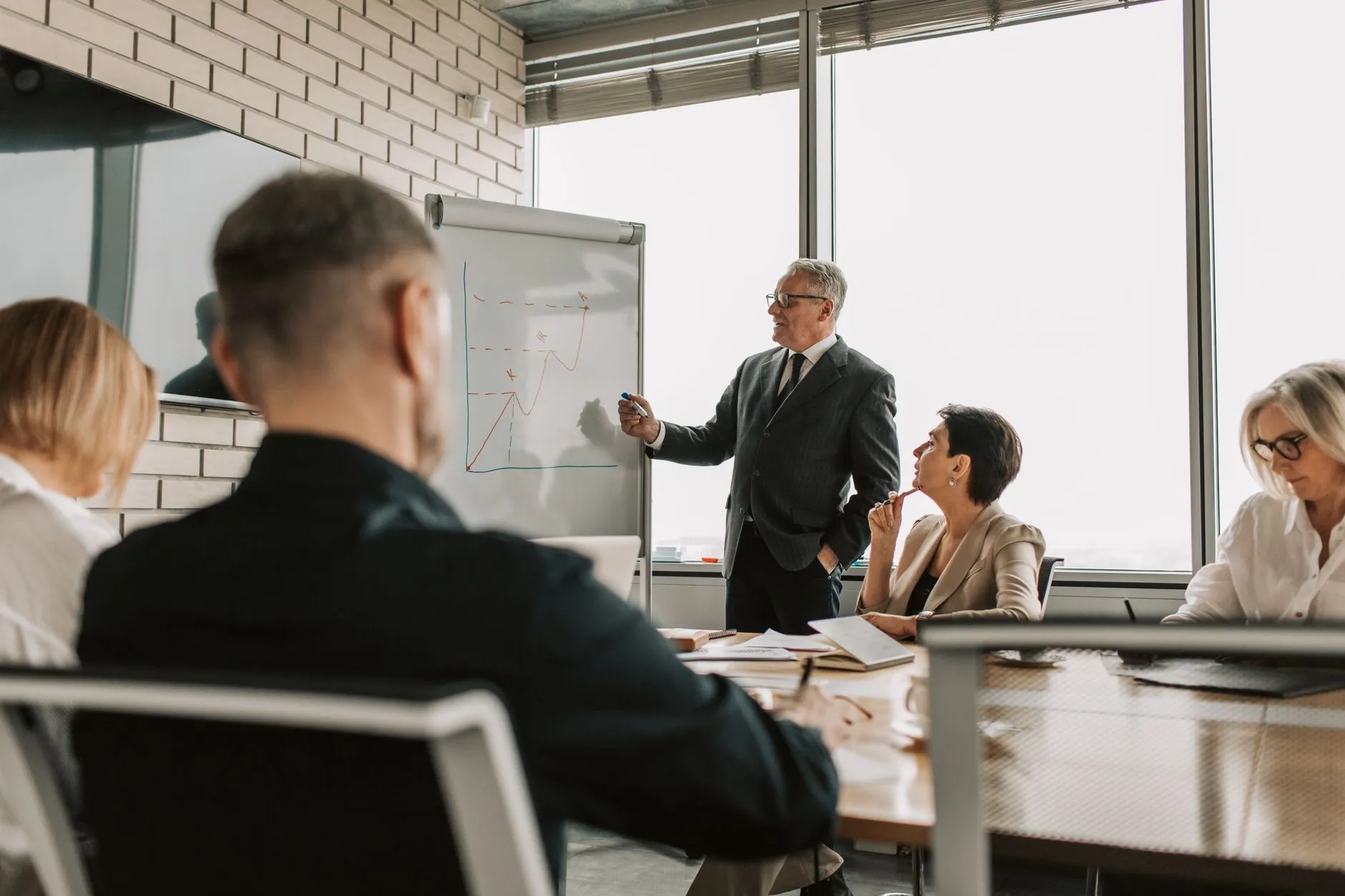 A senior executive in a suit presents a hand-drawn growth chart on a flip chart to a leadership team seated around a conference table.