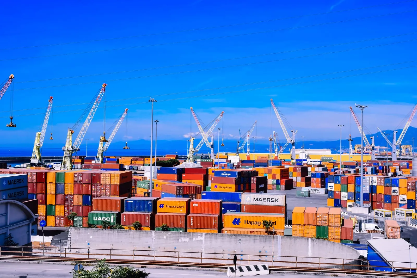 Container port under a bright blue sky with cranes towering over tall stacks of orange, red and blue shipping containers