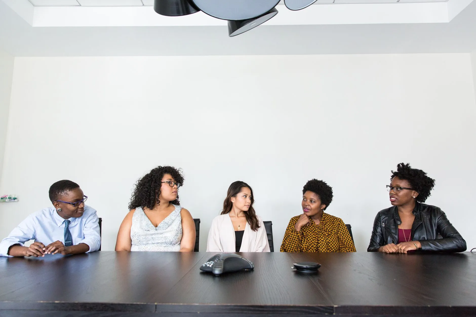 Five people sit in a row along a long dark conference table, glancing at one another in a plain white room