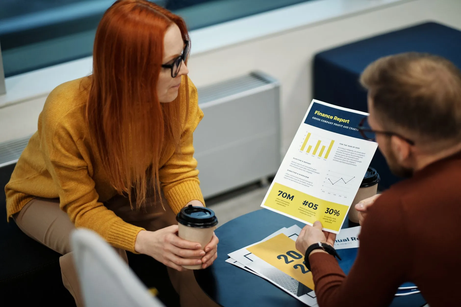 A woman with long red hair and glasses, holding a takeaway coffee cup, listens as a colleague holds up a printed finance report with bar and line charts at a small round table.