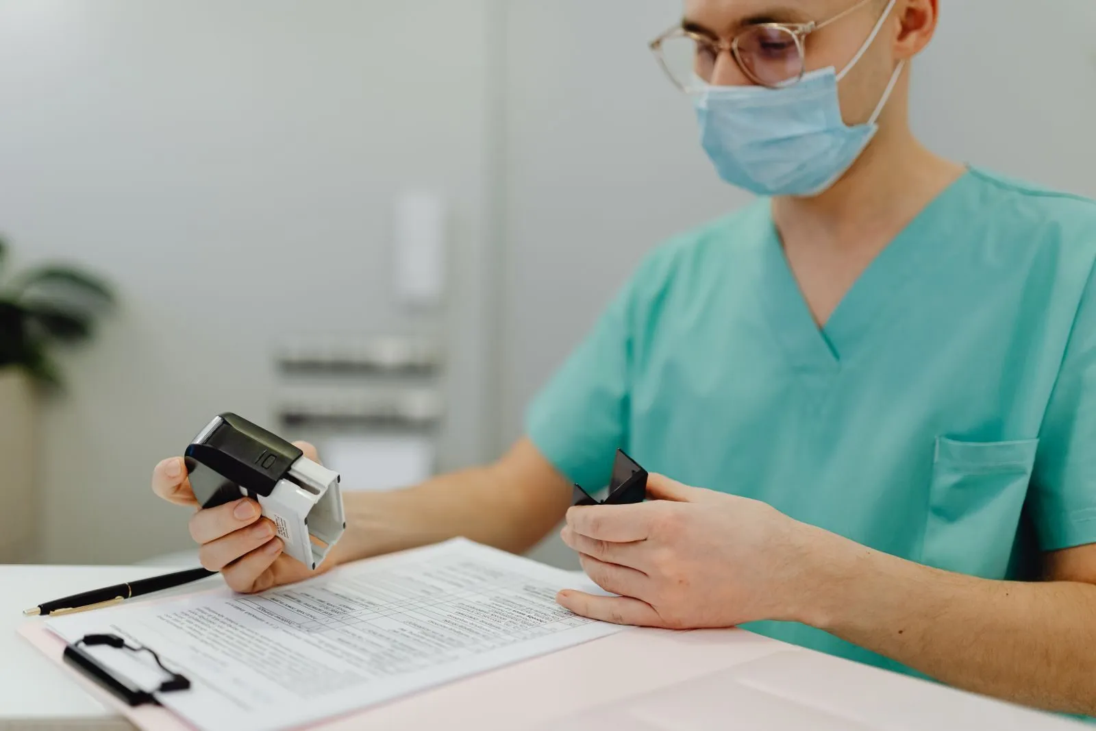 A clinic worker in teal scrubs and a surgical mask holds a self-inking stamp over a patient intake form on a white counter.