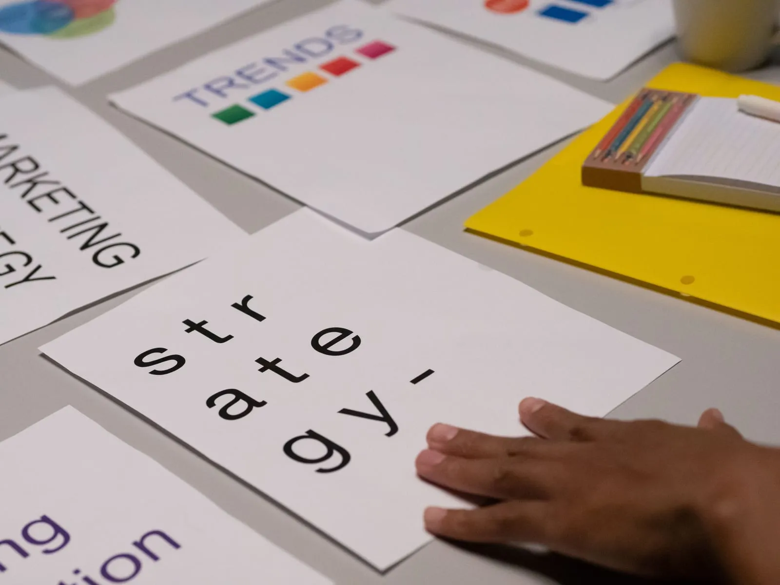 Hand rests on printed sheets spread across a table, with words like trends and strategy, beside a yellow folder and notepad