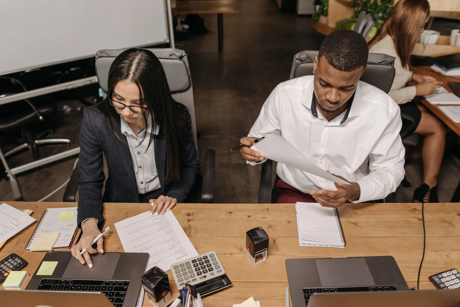 Seen from above, two young professionals work side by side at a wooden desk: a woman in glasses writes beside her laptop and a printed balance sheet, while a man in a white shirt reads a page he holds up, with calculators and a desk stamp between them.
