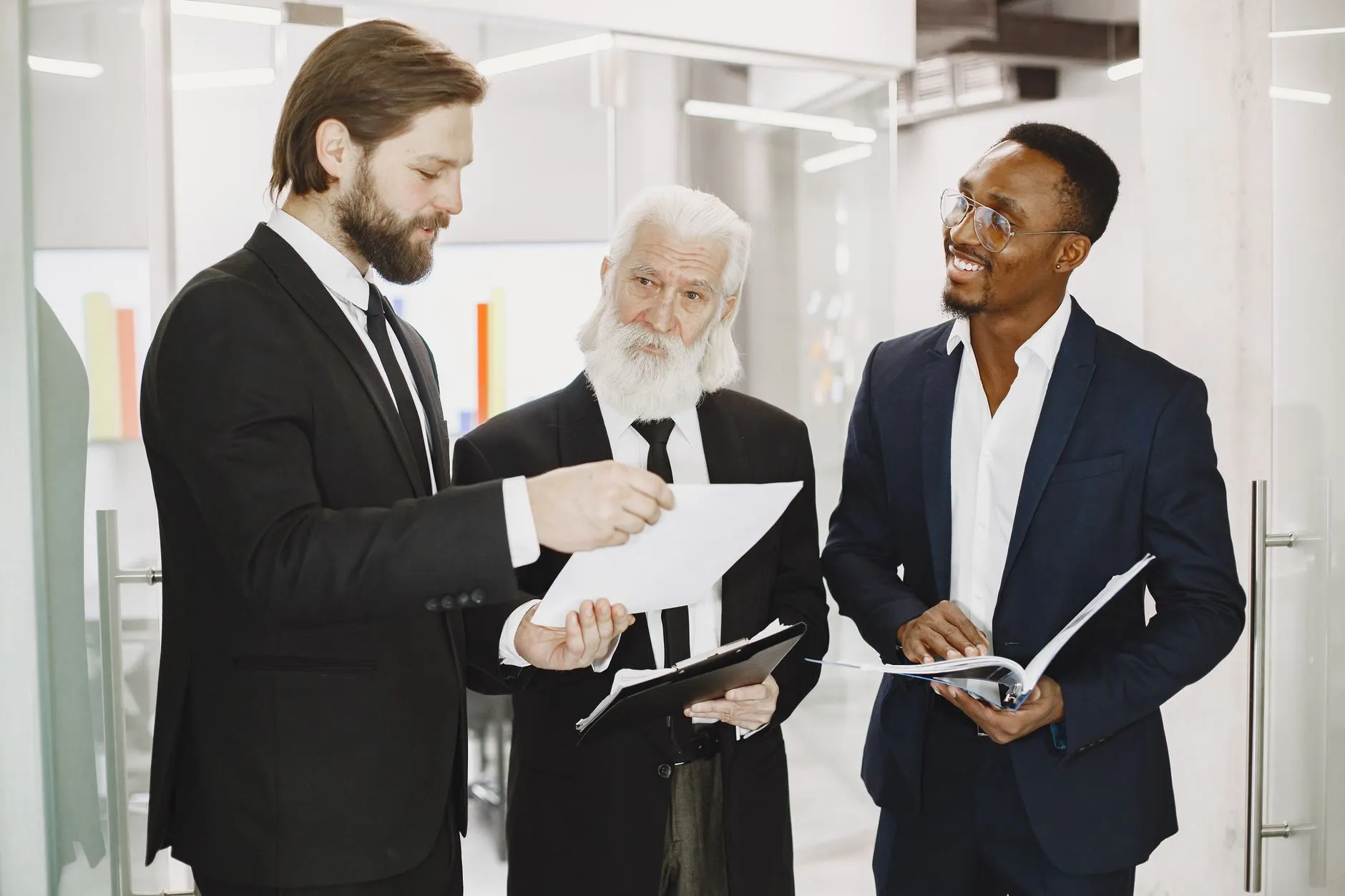 Three business professionals in suits reviewing printed documents together at an office table
