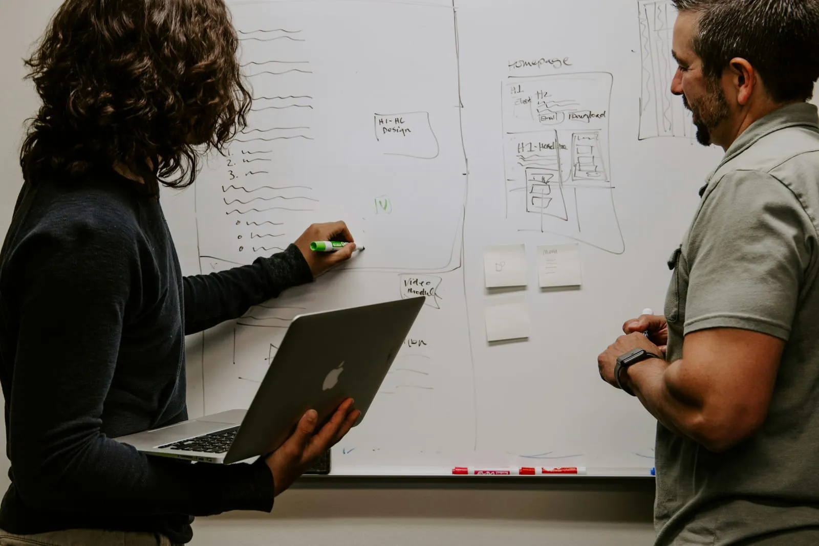 Two colleagues sketch a website layout on a whiteboard, one drawing with a green marker while holding a laptop and the other watching.