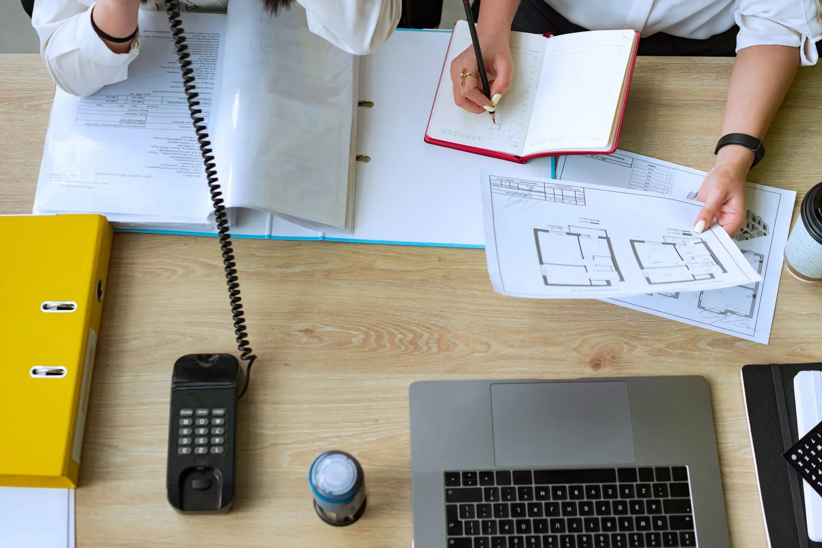 Overhead view of two people at a wooden desk, one taking notes in a red notebook and one holding floor plans, beside a laptop, desk phone and binders.