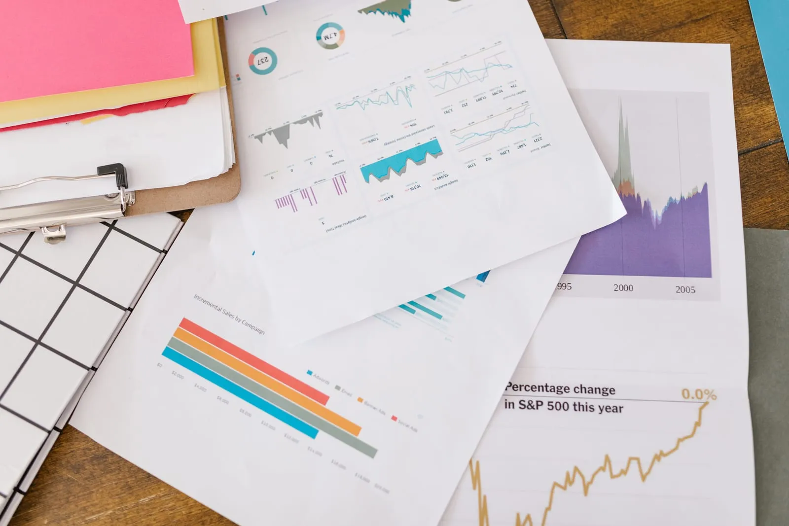 Printed charts and graphs, including bar, line, donut and stock market plots, spread across a wooden desk next to a clipboard and colored folders.