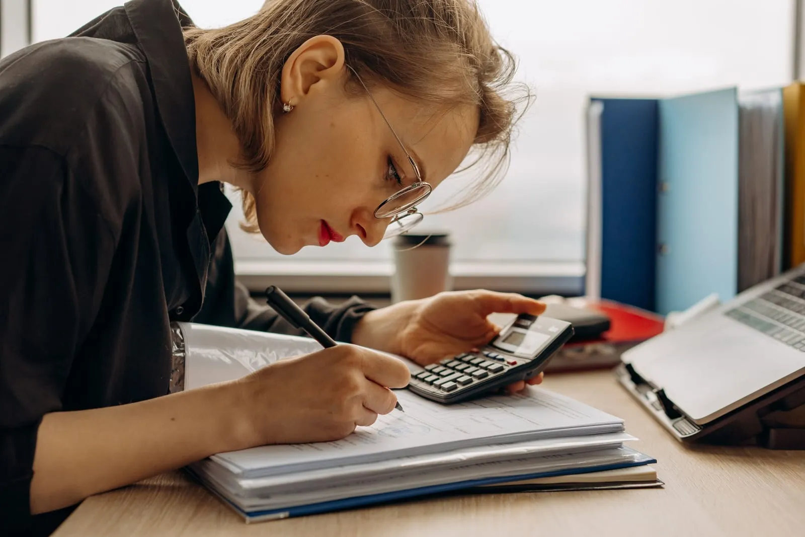 A woman with glasses leans over a stack of paperwork, writing with a pen while holding a desk calculator, with binders and a laptop beside her.