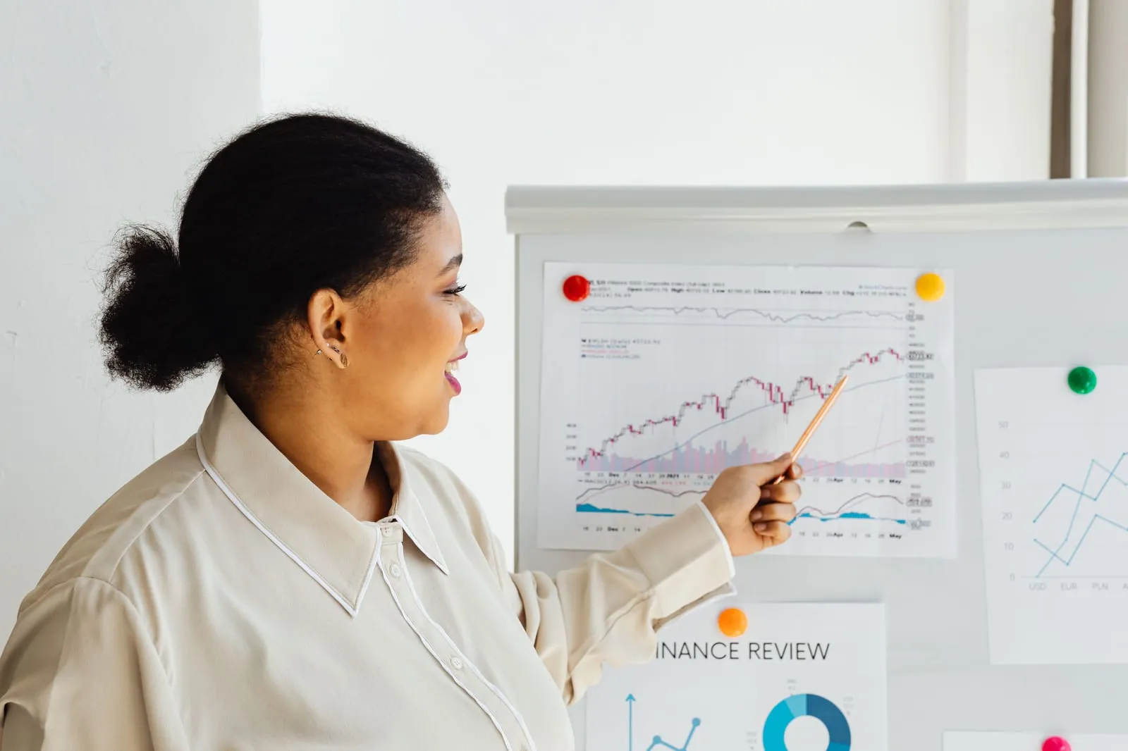 Smiling woman points a pen at printed stock charts and a finance review sheet pinned to a whiteboard