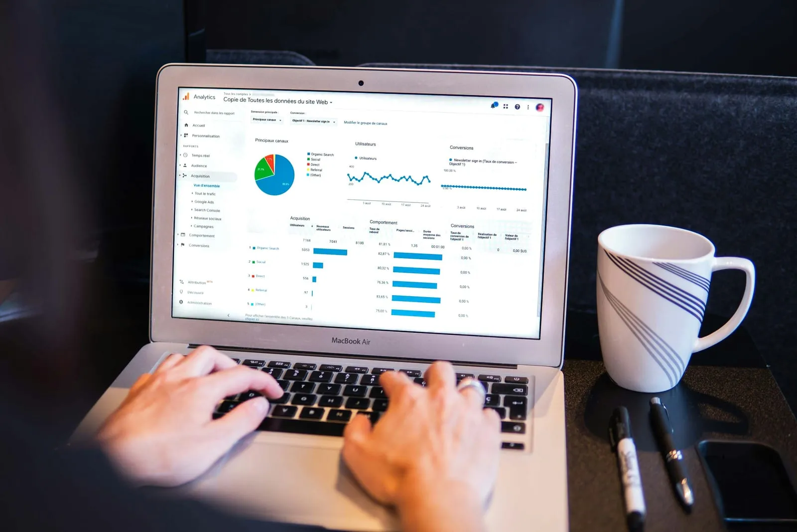 Hands typing on a laptop that shows a web analytics dashboard with a pie chart and line graphs, a white mug and pens beside it