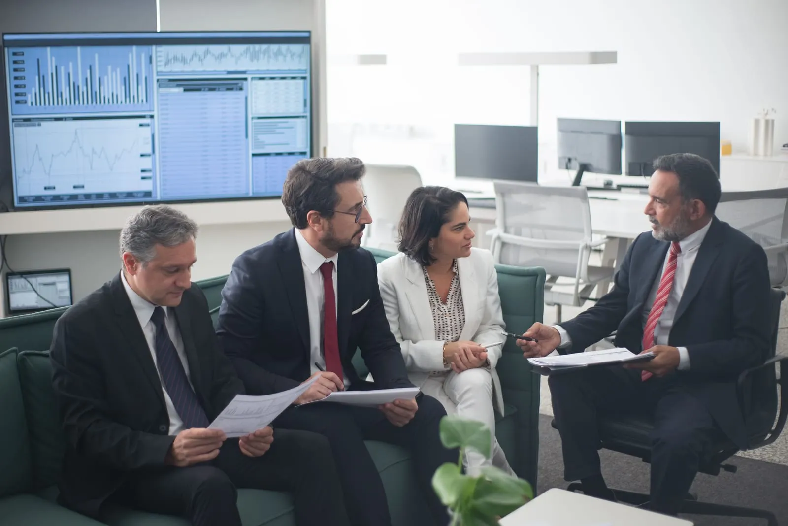 Four business colleagues in suits sit on a sofa reviewing printed reports and talking, with a large data display on the wall behind them