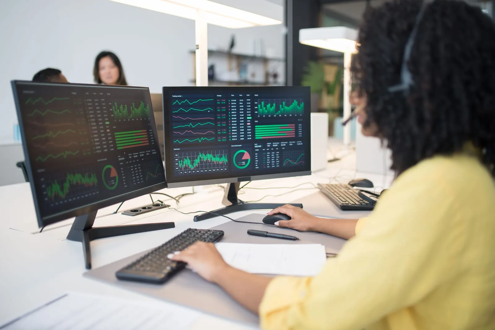 Woman wearing a headset works at two monitors filled with green trading charts in a bright open-plan office