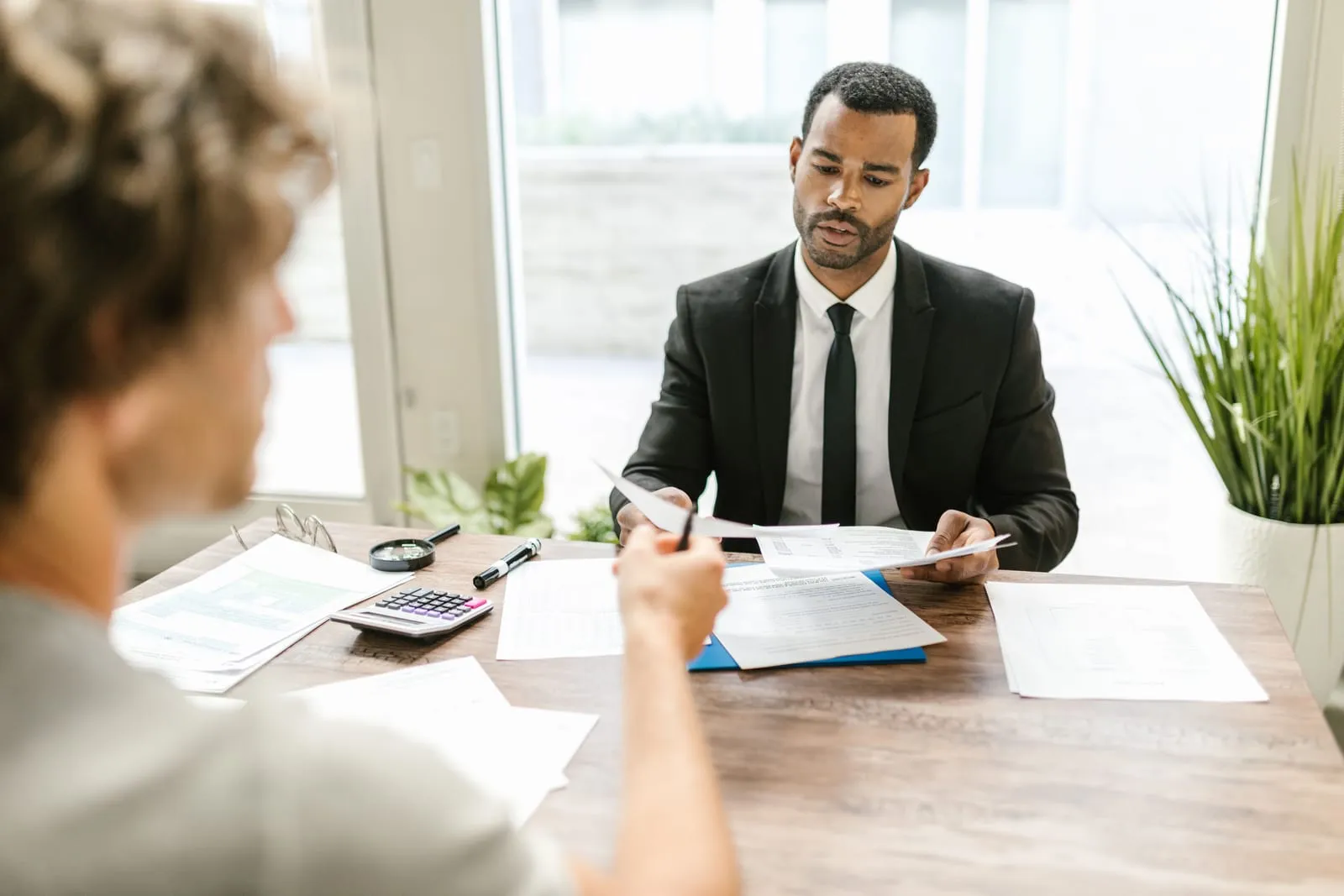 Man in a dark suit reviews papers across a wooden desk while a client in the foreground hands him a document, a calculator nearby