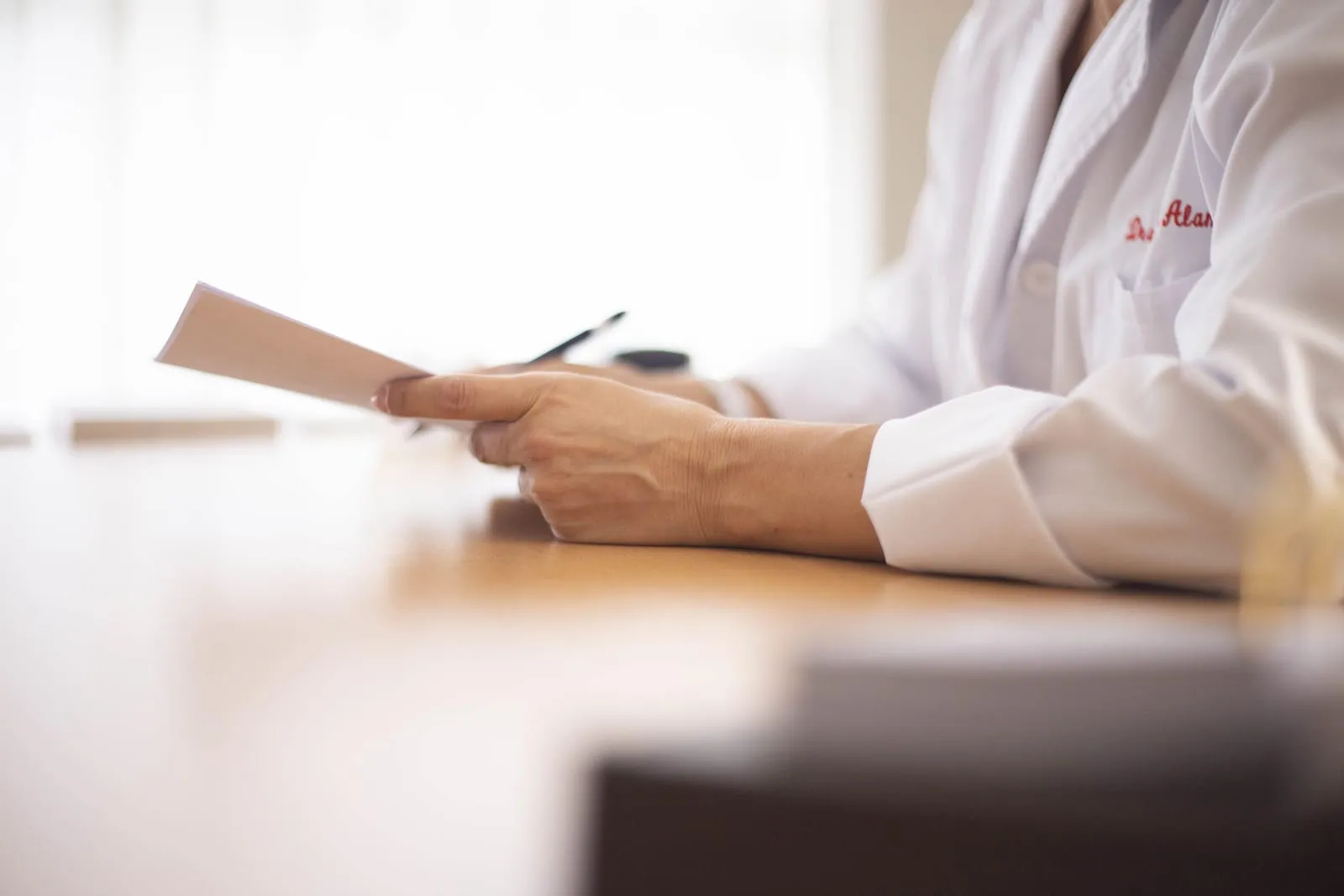 Person in a white lab coat sits at a wooden desk holding a few sheets of paper and a pen, shot close to table level