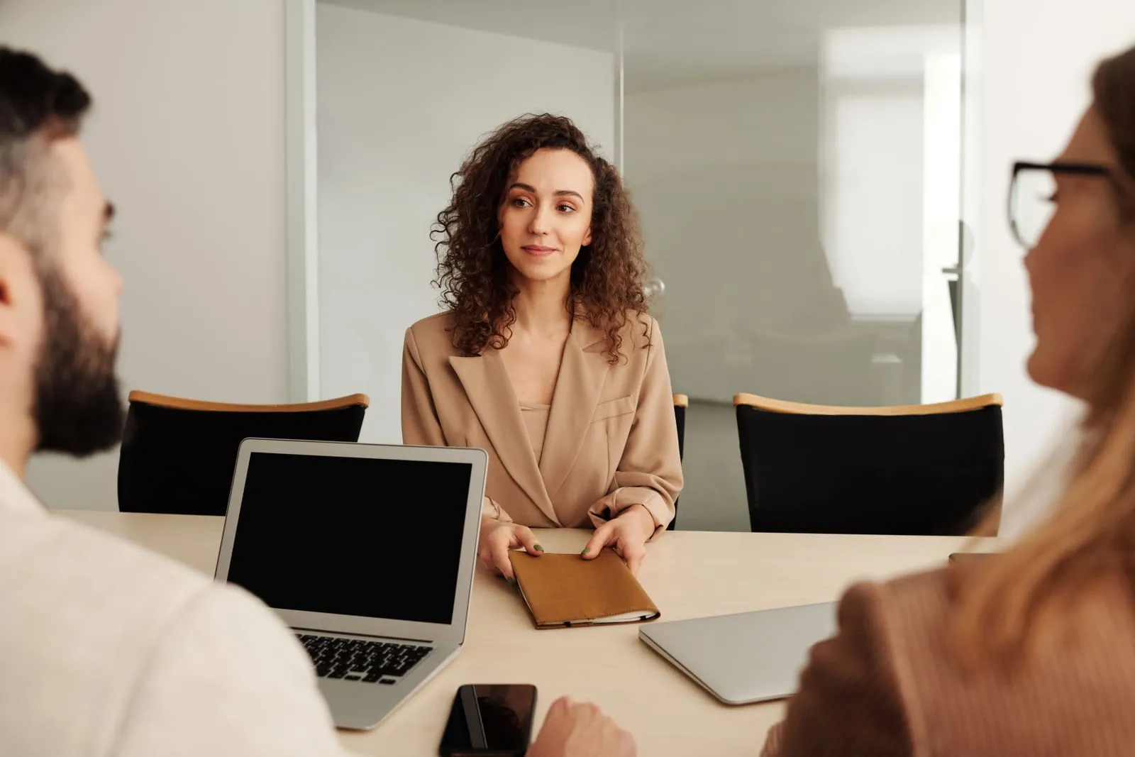 Candidate in a beige blazer holds a notebook at a conference table while two interviewers with a laptop face her