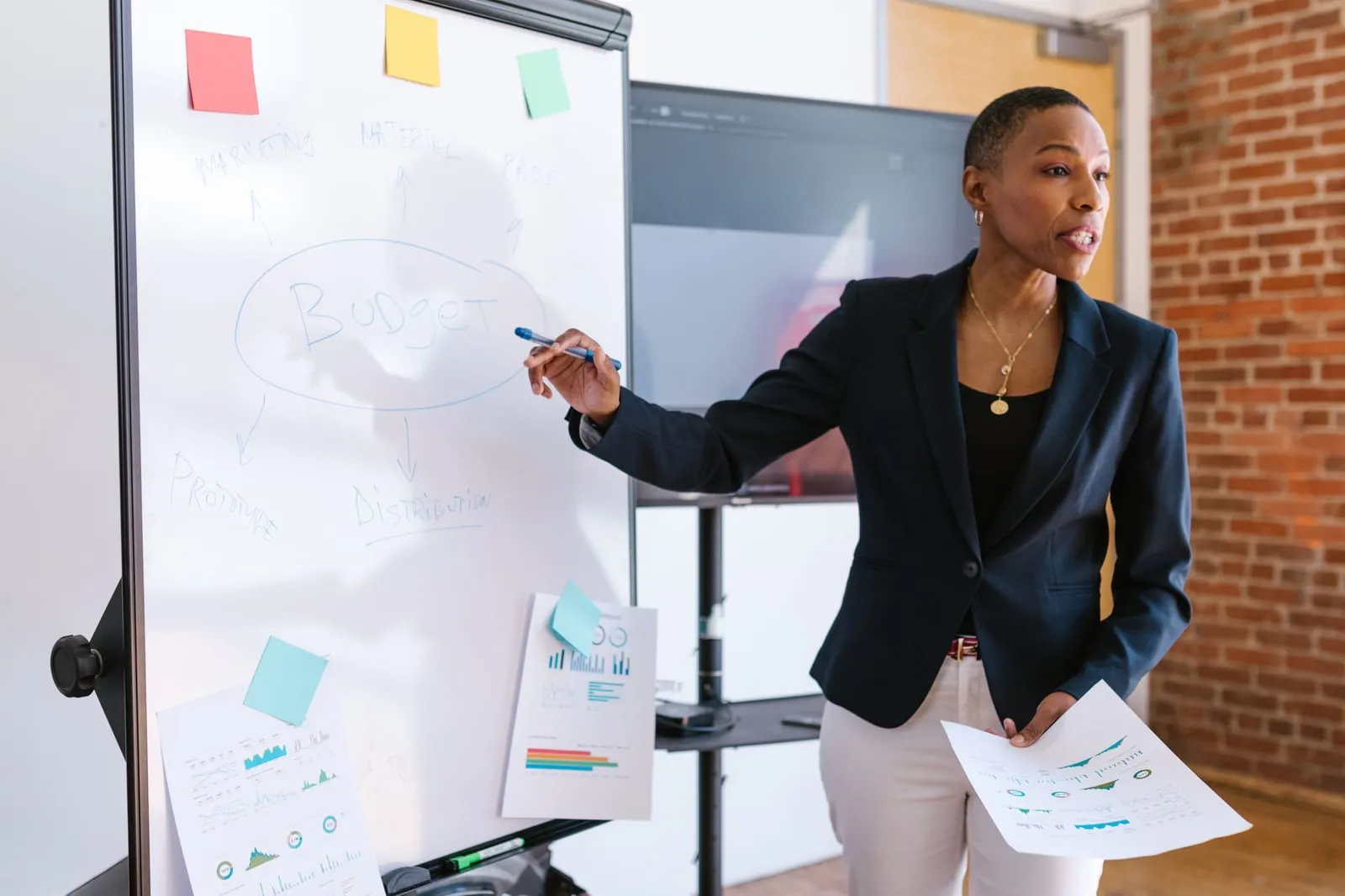 Woman in a navy blazer points to the word budget on a whiteboard diagram while holding a printed chart in her other hand