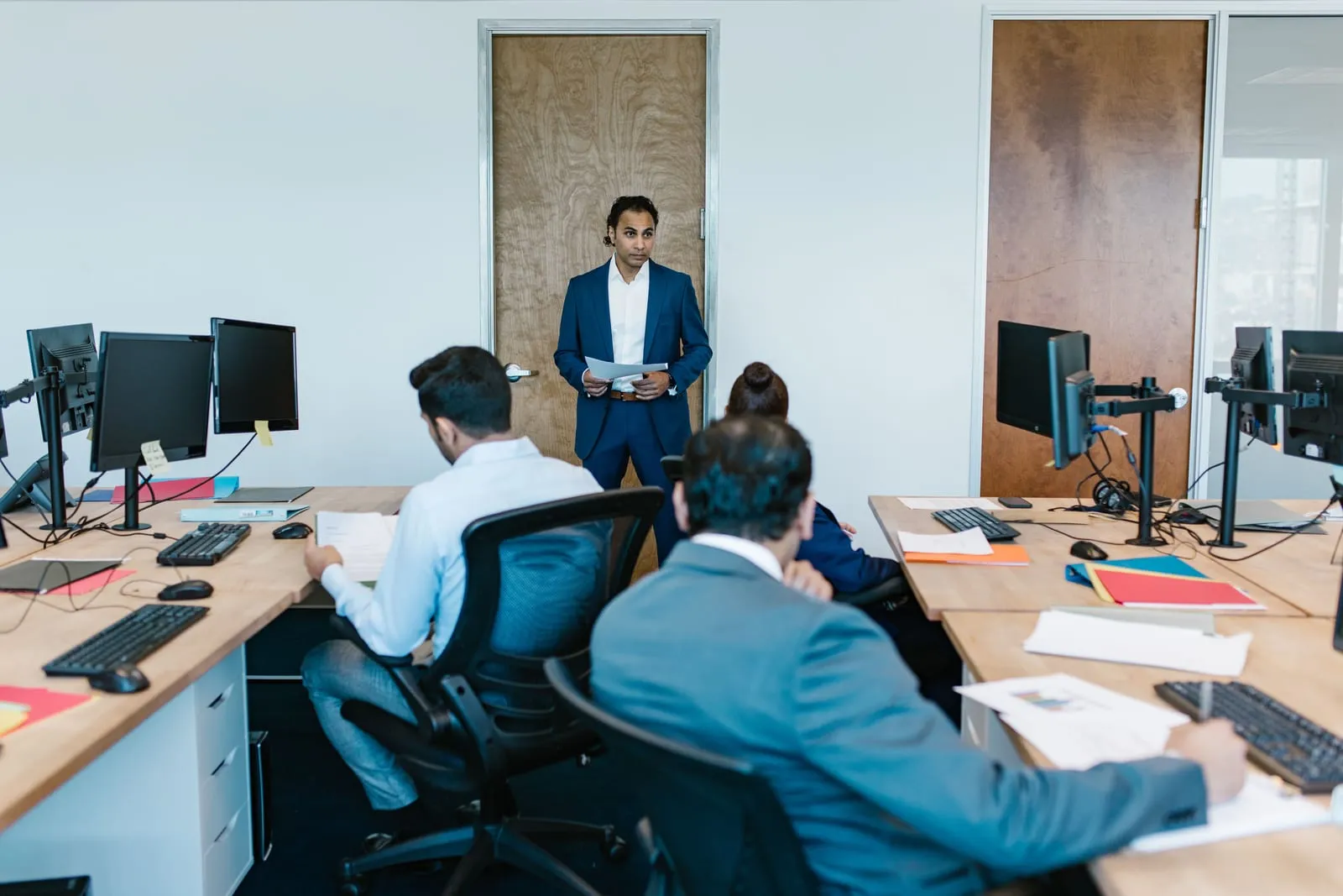 Manager in a blue suit stands at the front of an open office holding papers while seated employees work at desks with monitors