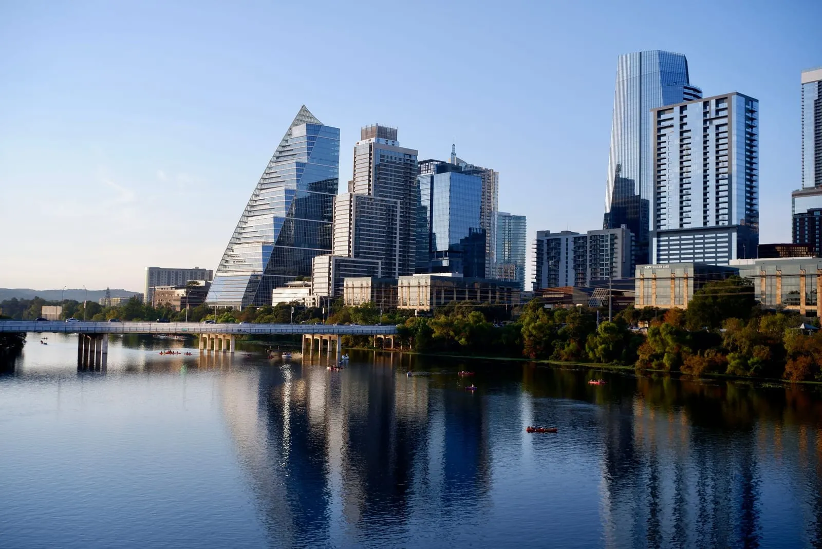 Glass towers, one shaped like a steep wedge, reflected in a calm river below a road bridge