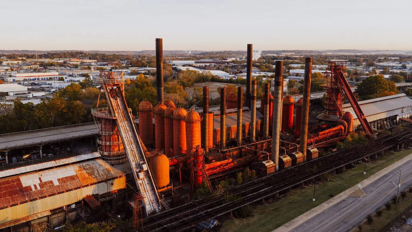 The rusted smokestacks and furnaces of an old ironworks lit by sunset, the city spreading out behind
