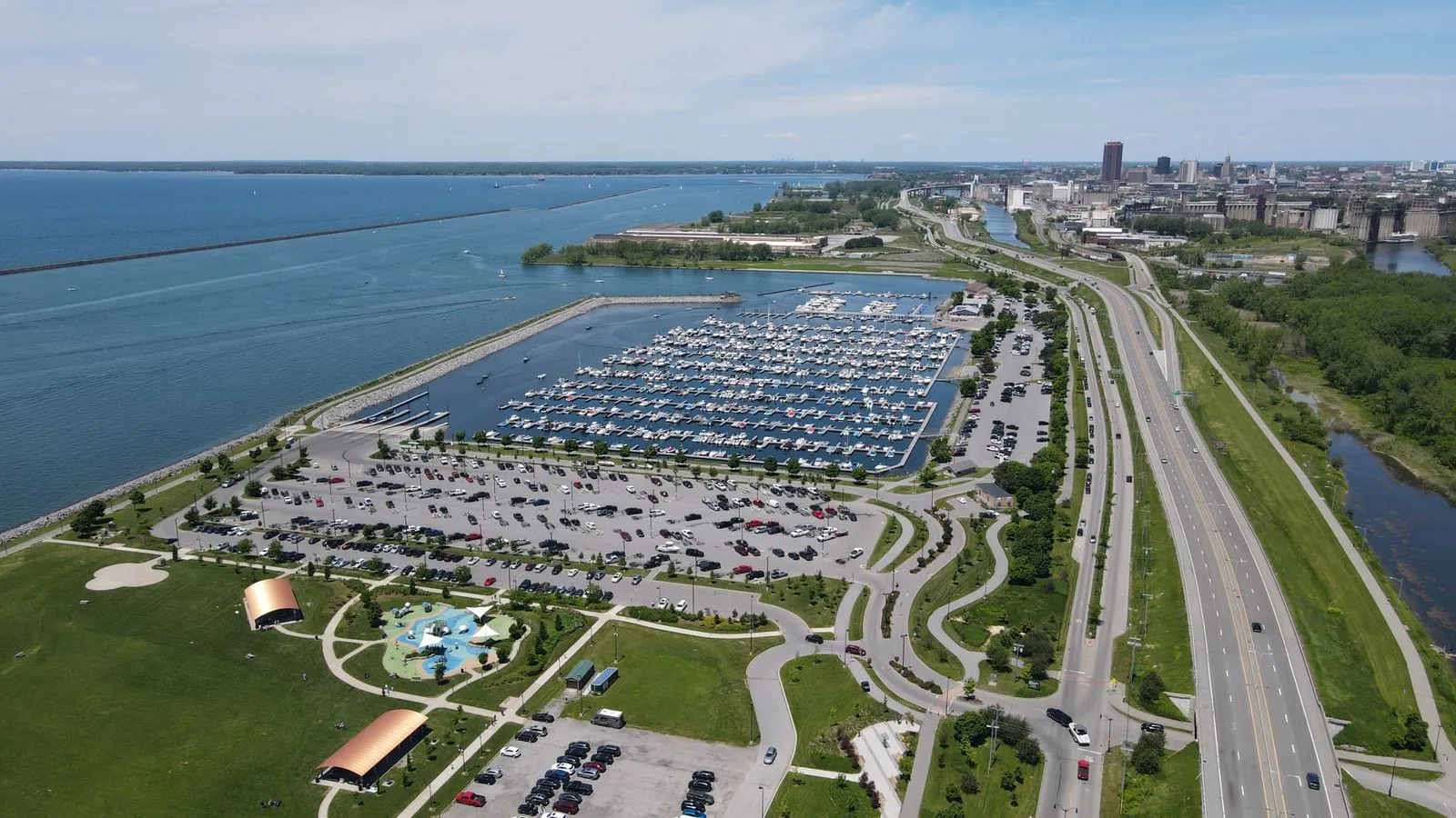 Aerial view of a lakefront marina and park beside a highway, a downtown skyline in the distance