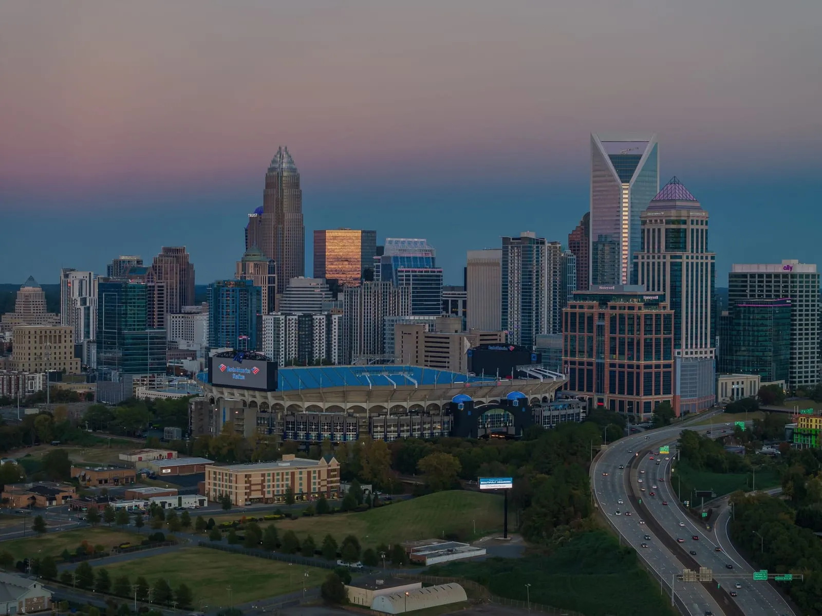 A downtown skyline at dusk with a stadium in front and a highway curving past