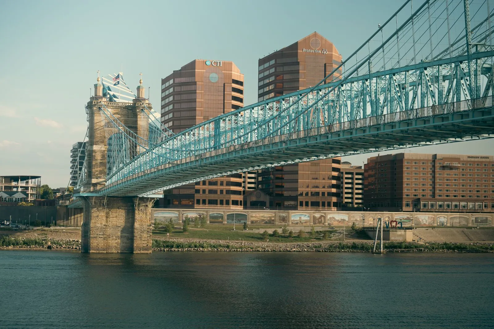 A blue steel suspension bridge with stone towers crossing a river in front of downtown office buildings