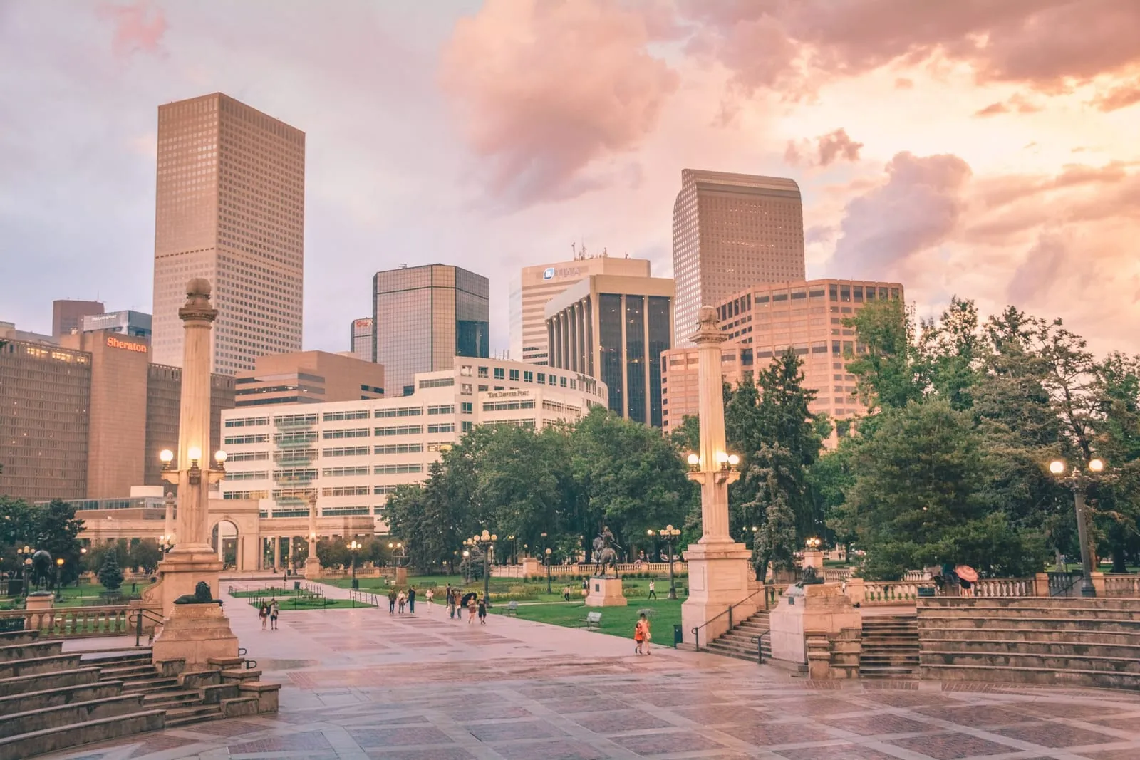 A paved plaza and lawn with ornamental lamps leading toward downtown office towers at dusk