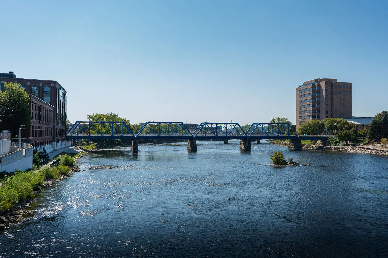 A blue steel truss footbridge crossing a wide river, city buildings on the far bank