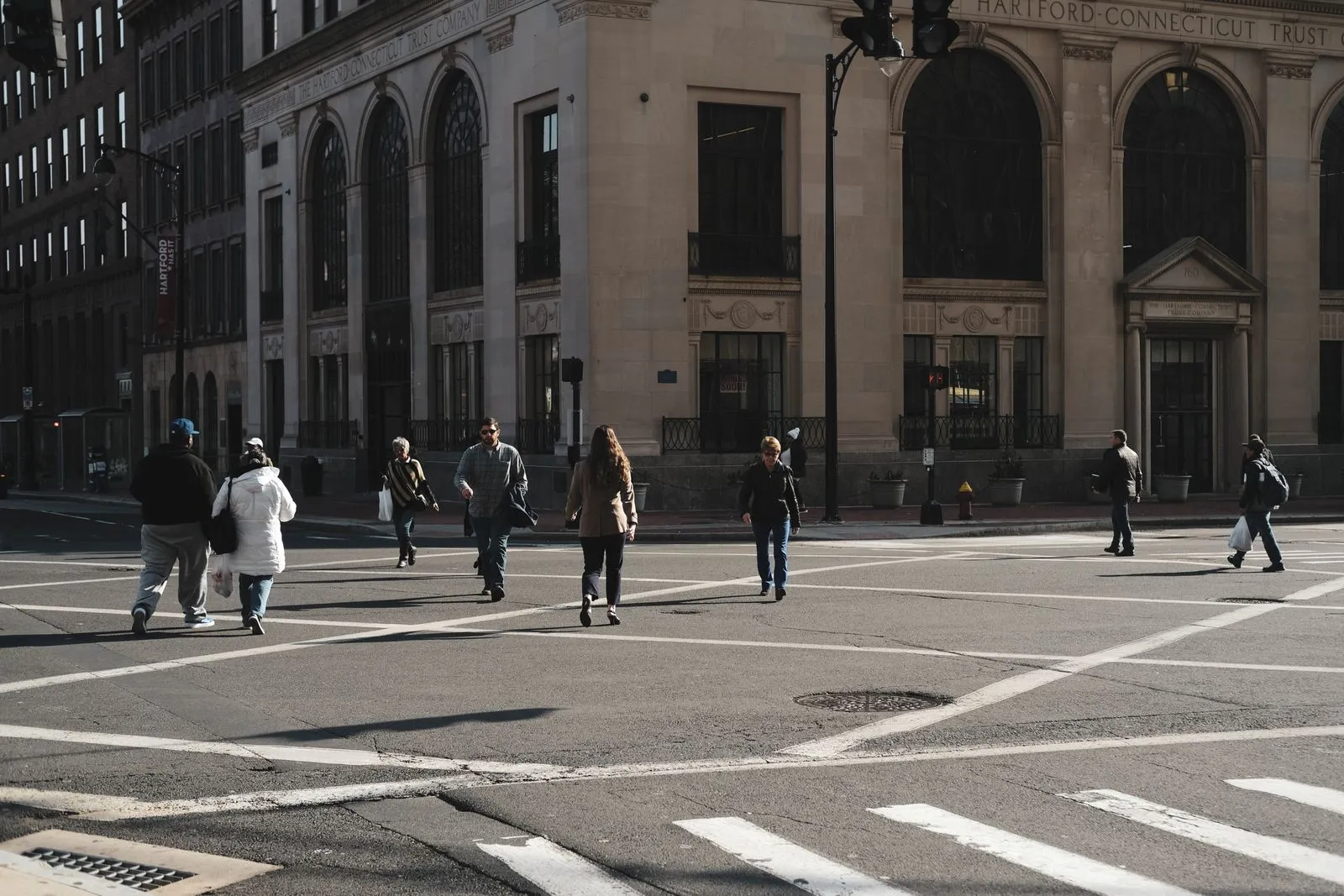 People crossing a wide downtown intersection in front of an arched stone office building