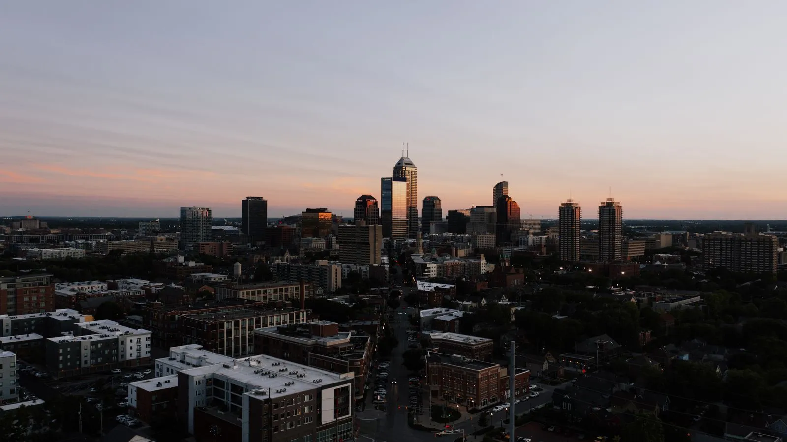 A downtown skyline at dusk seen from above, low brick blocks and trees in the foreground