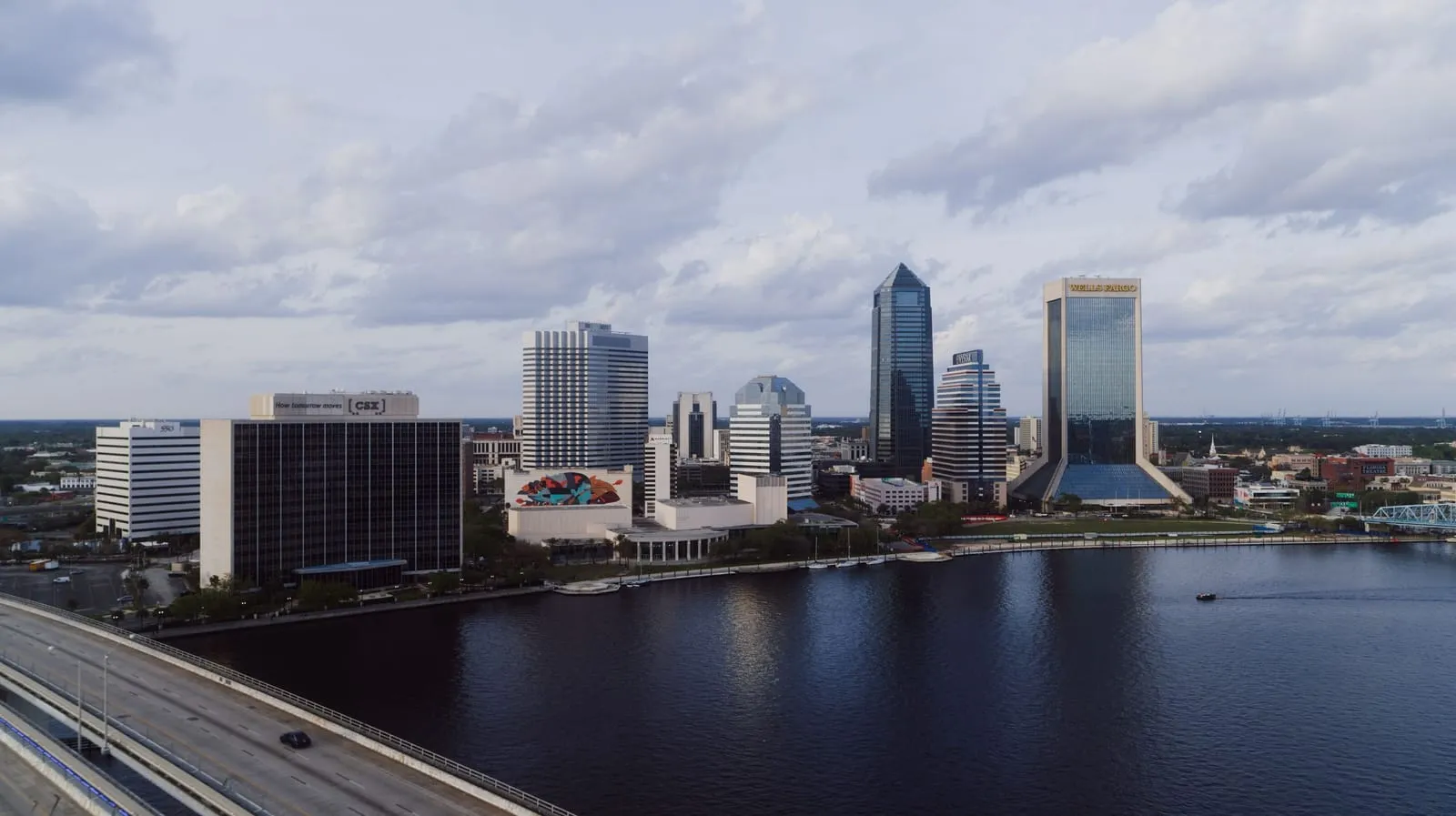A downtown skyline across a wide dark river under scattered clouds, a highway in the corner