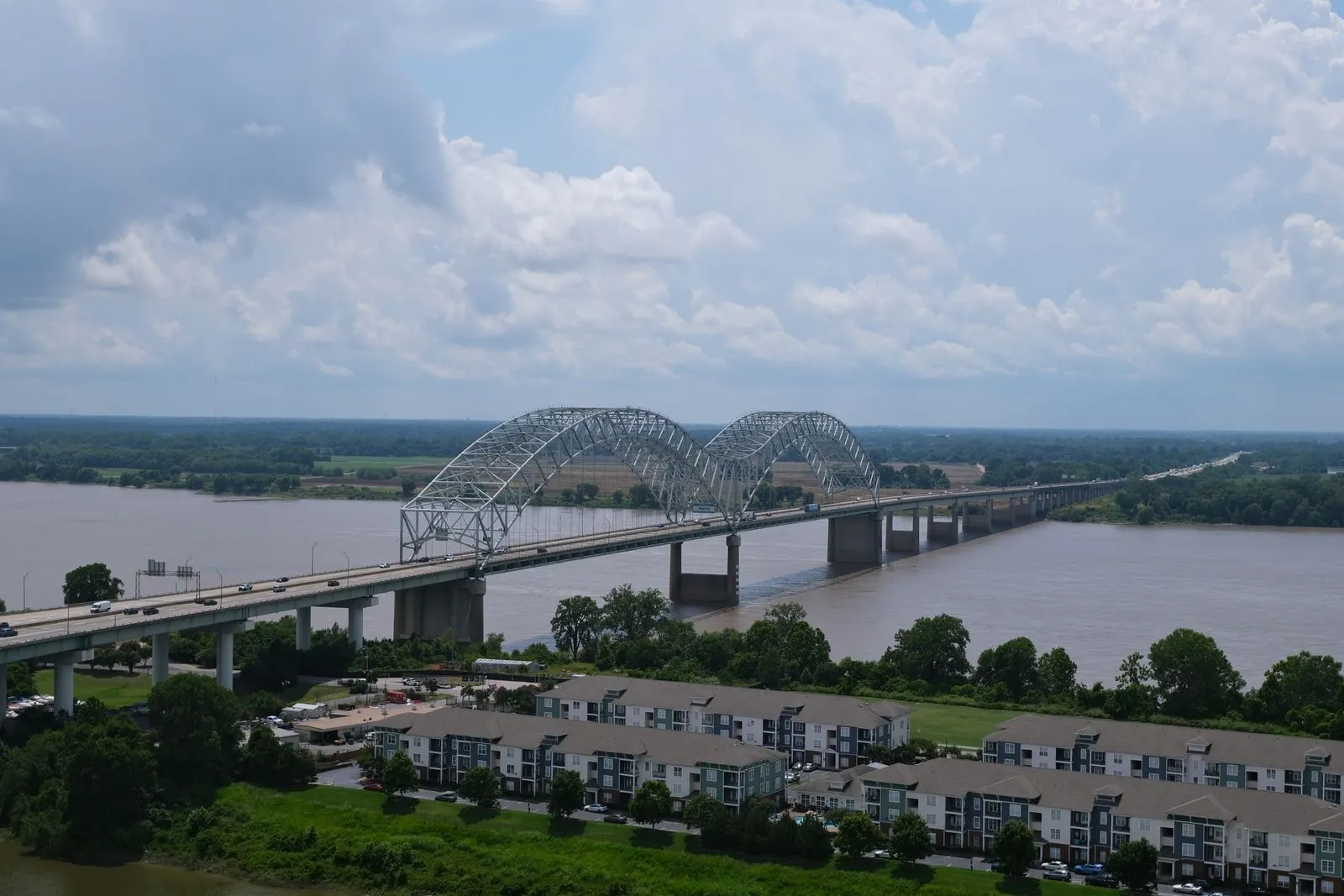 A double-arched steel bridge carrying a highway across a wide brown river, apartment blocks and trees in the foreground