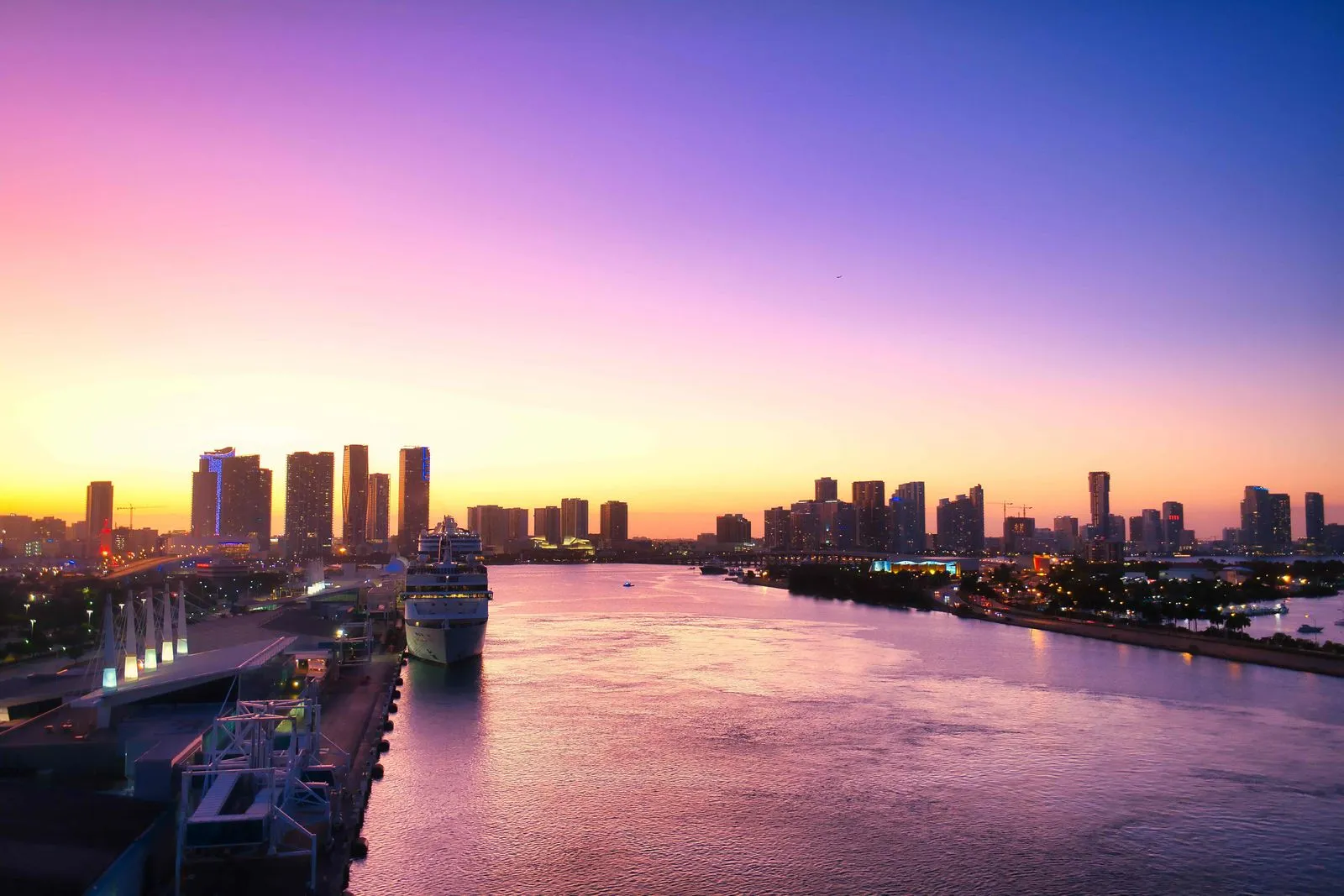 A waterway glowing pink at sunset, a cruise ship docked beside a line of high-rise towers