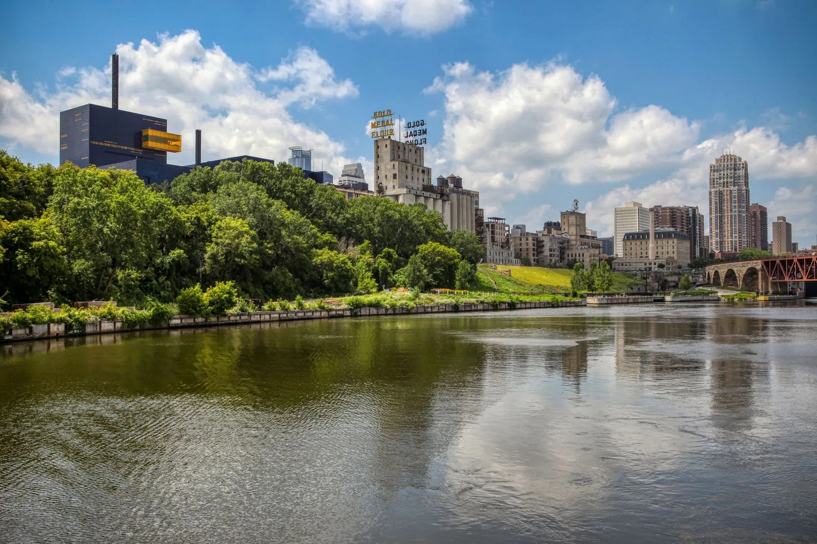 A downtown skyline beyond a calm river with green banks under puffy clouds