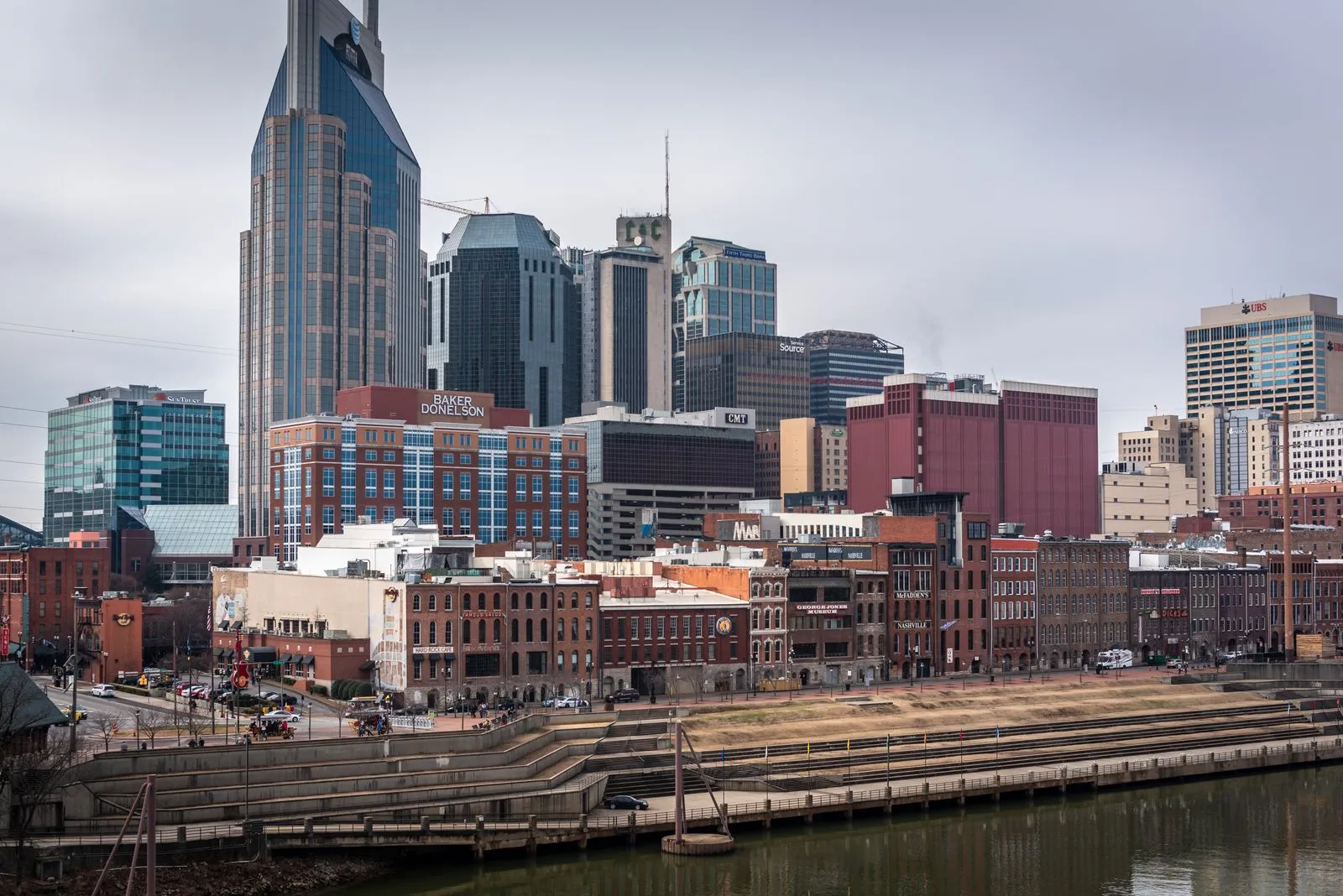 A downtown skyline with a tall twin-spired tower above brick riverfront buildings and wide steps down to a river