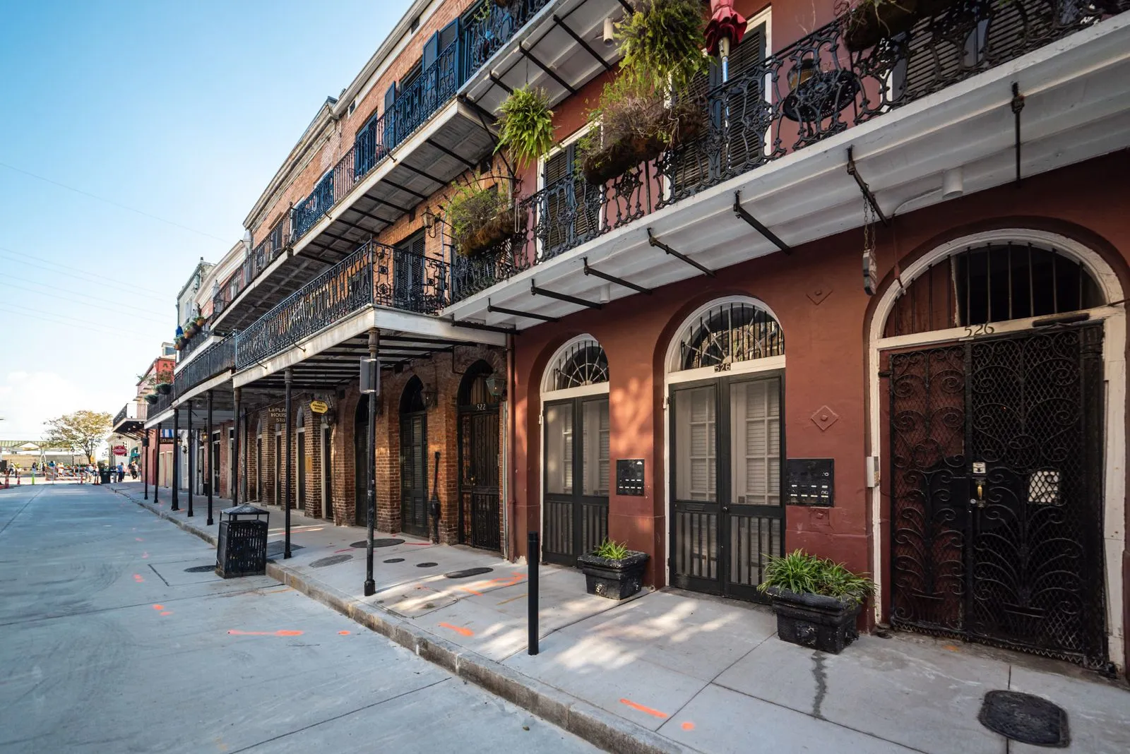 A red brick building with arched doorways and wrought-iron balconies hung with plants along a quiet street